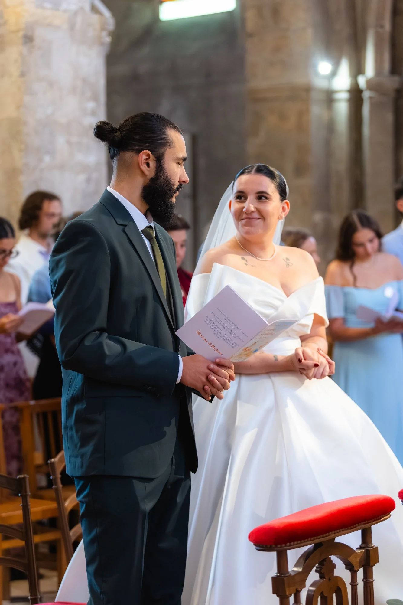 Un couple lors de leur mariage, le homme porte un costume noir avec une cravate et une barbe, la femme porte une robe de mariée blanche avec un collier et un tatouage visible sur l'épaule, ils se regardent avec amour dans une église avec des invités 