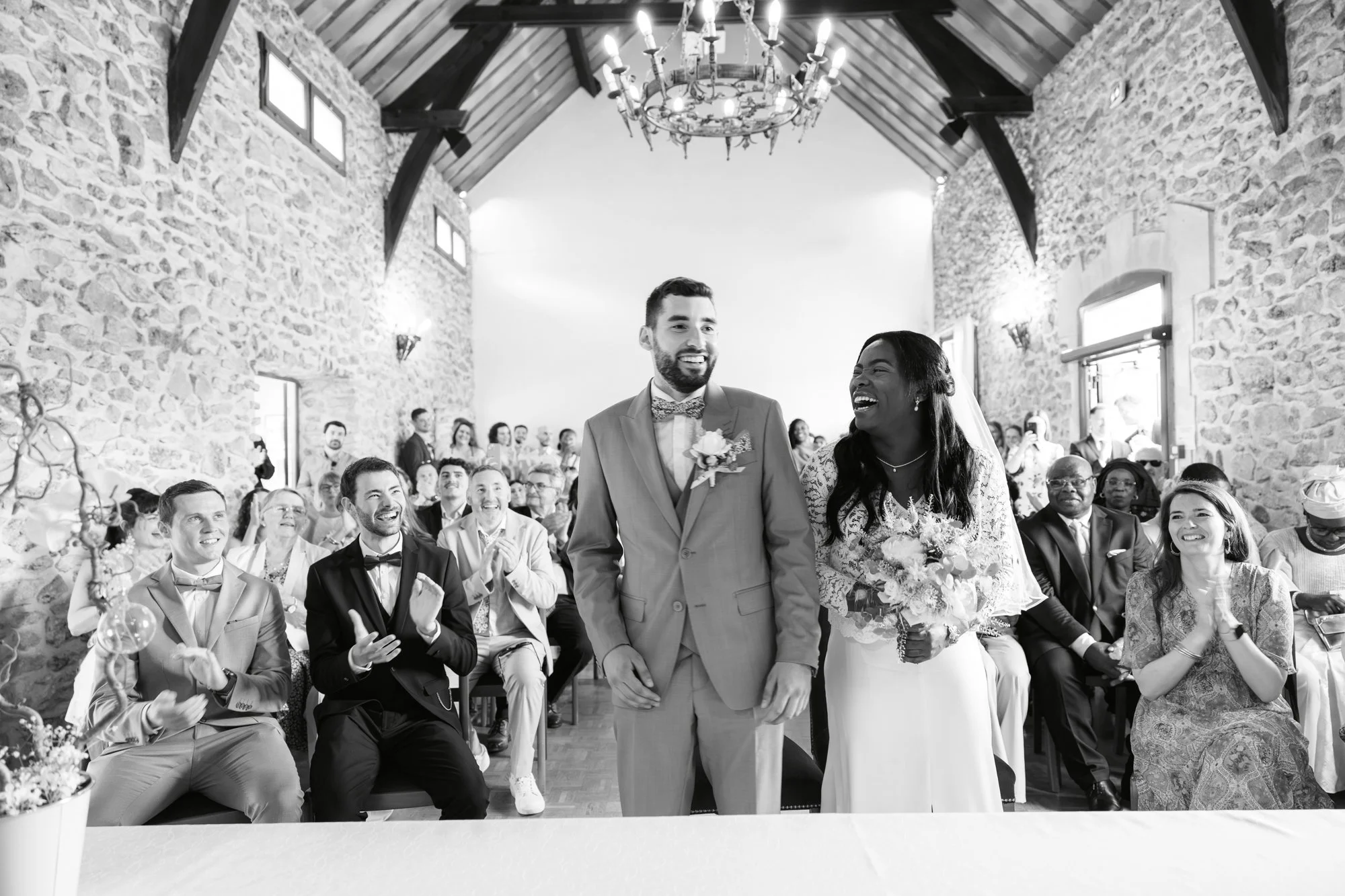 Un couple de mariés lors d'une cérémonie dans une salle avec murs en pierre et plafond en bois, entouré d'invités assis qui applaudissent et sourient.