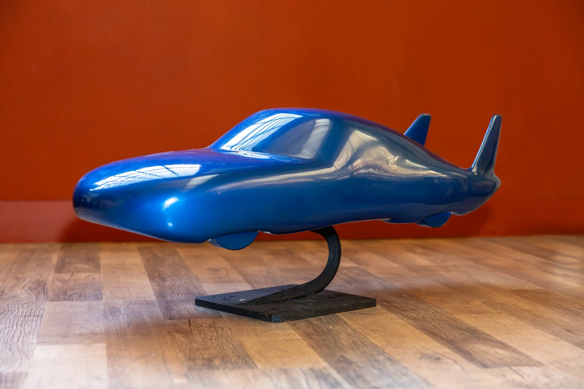 Une maquette d'avion futuriste de couleur bleue, exposée sur un socle noir, posée sur une table en bois. L'arrière-plan est orange.