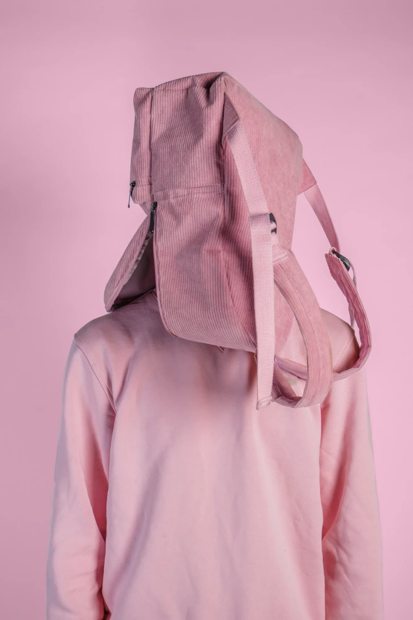 Personne portant un hoodie rose avec un sac à dos rose clair, contre un fond rose pâle.