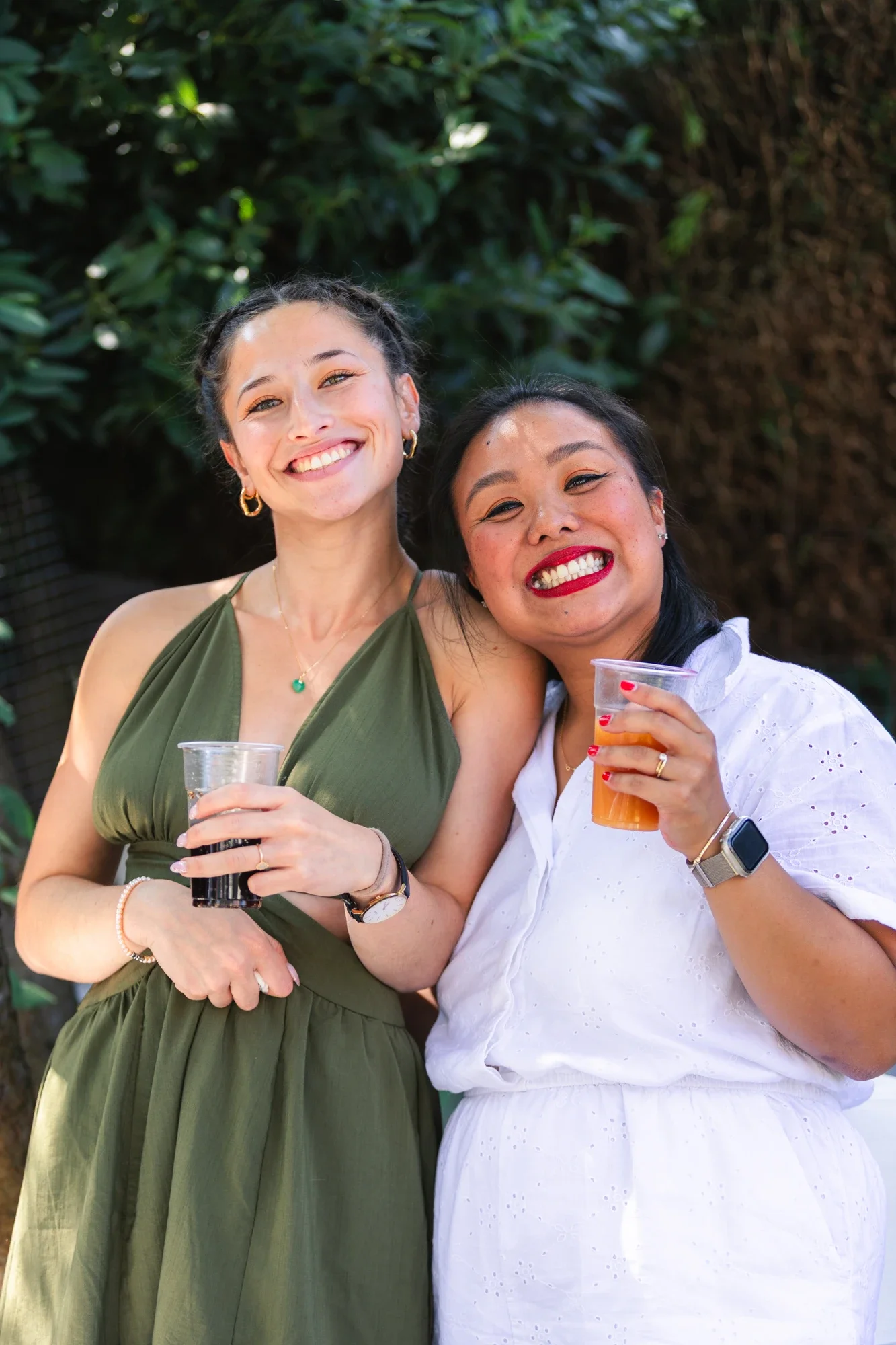 Deux femmes souriantes tiennent des verres de boisson lors d'une fête en extérieur, entourées de végétation.