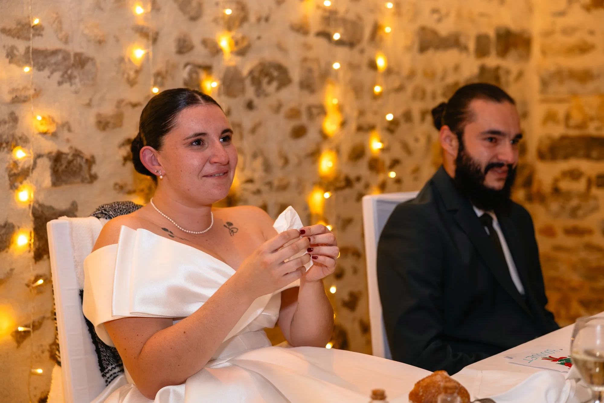 Une femme habillée en robe de mariage blanche, assise à une table lors d'une réception, tenant un mouchoir, avec un mur en pierre en arrière-plan décoré de guirlandes lumineuses. À côté, un homme en costume noir avec une barbe.