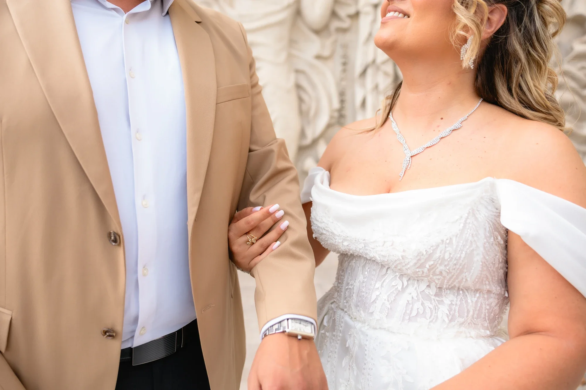 Un couple marié lors d'une cérémonie, la femme porte une robe de mariée blanche et un collier élégant, et l'homme porte un costume beige avec une chemise blanche. L'homme tient la main de la femme, qui affiche un sourire.