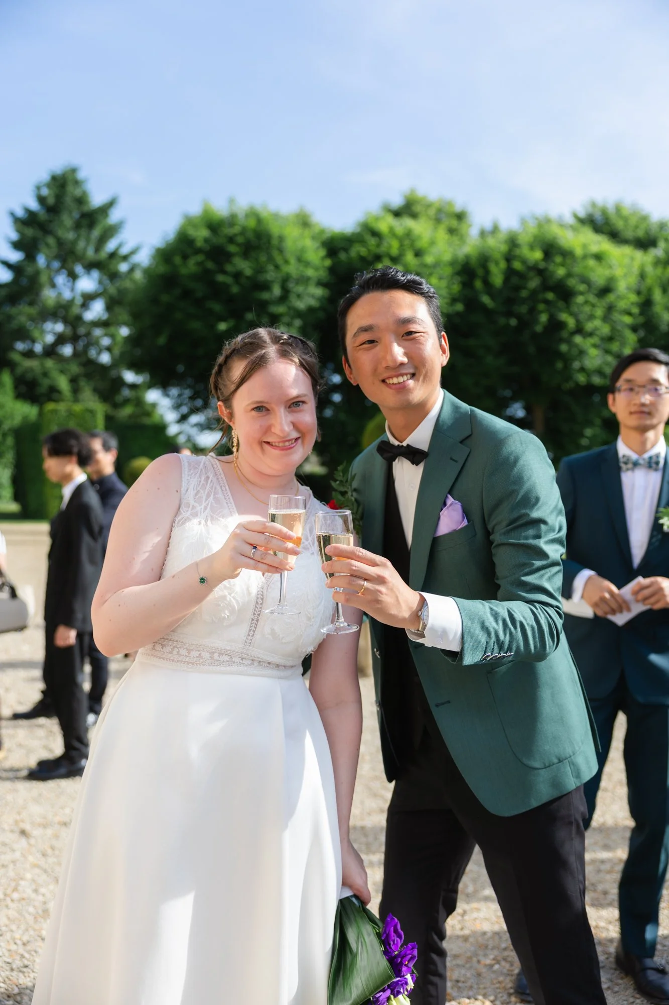 Un couple en robe de mariage et costume tenant des flûtes de champagne lors d'une célébration en plein air, avec un groupe d'invités en arrière-plan, arbres verts et ciel bleu
