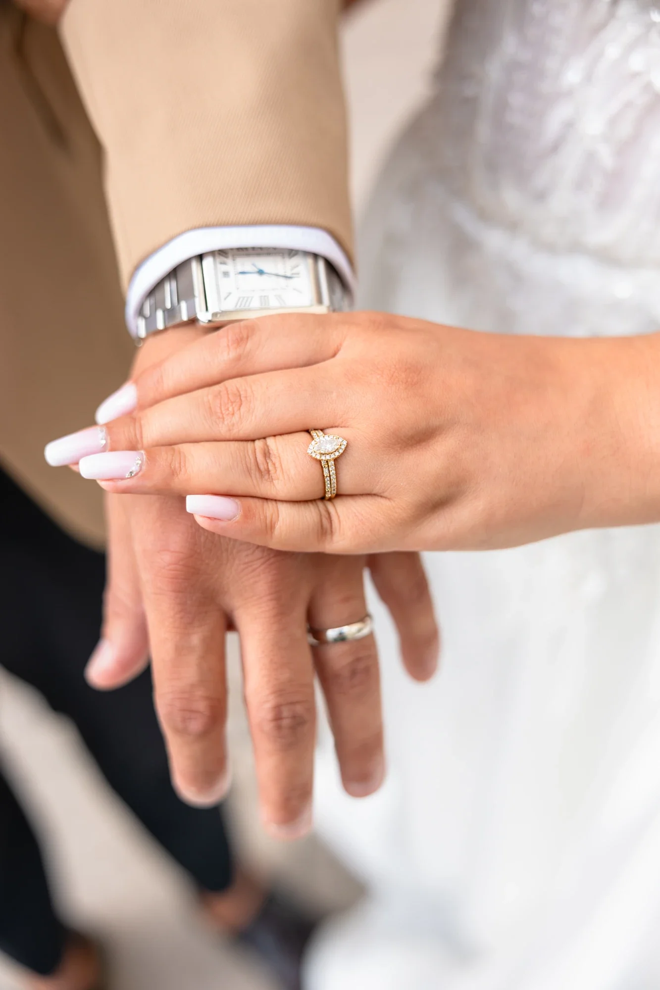 Une main portant une bague en or avec un diamant, une montre à cadran rectangulaire, et une autre main portant une bague de mariage. Le fond est flou.