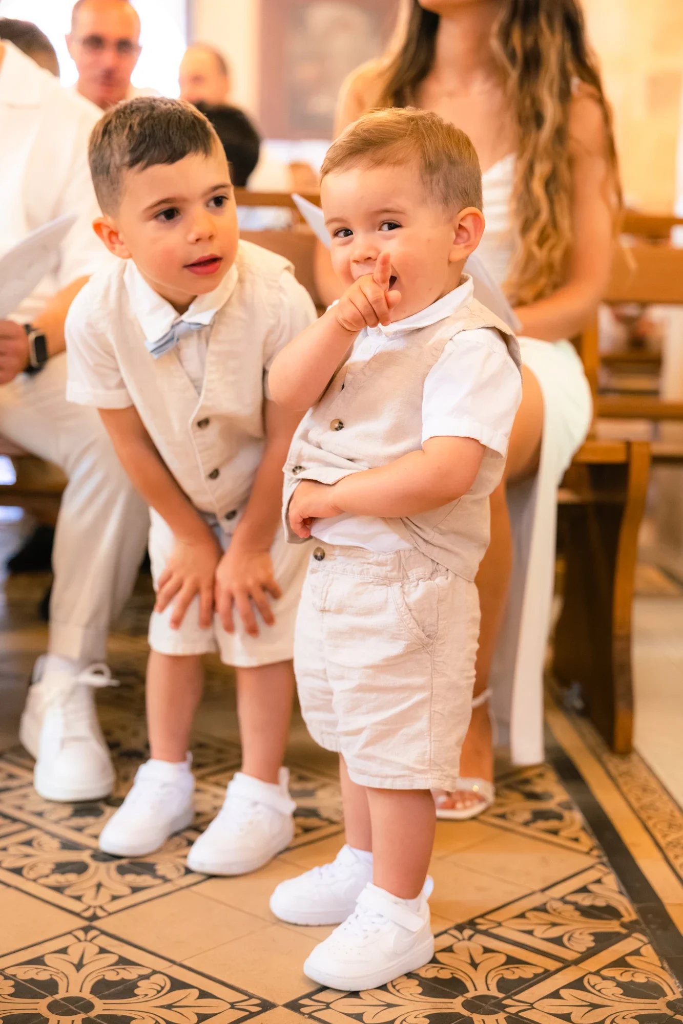 Deux jeunes garçons habillés en blanc avec des baskets blanches, debout sur un sol en carrelage décoratif, dans un espace intérieur, probablement lors d'une cérémonie ou événement.