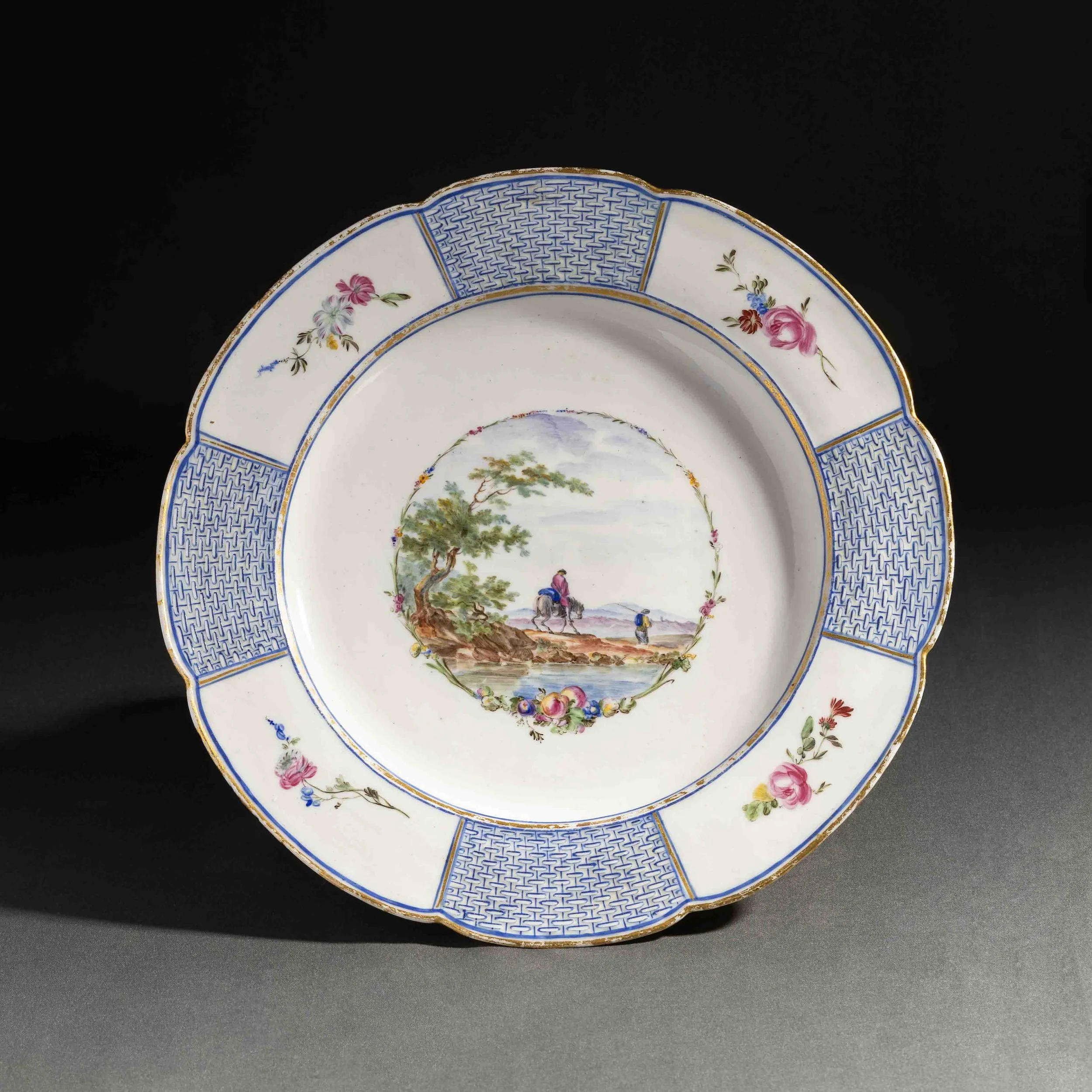 Une assiette en porcelaine décorée de motifs floraux, d'une scene pastorale avec des personnes à cheval et de motifs bleus et dorés.