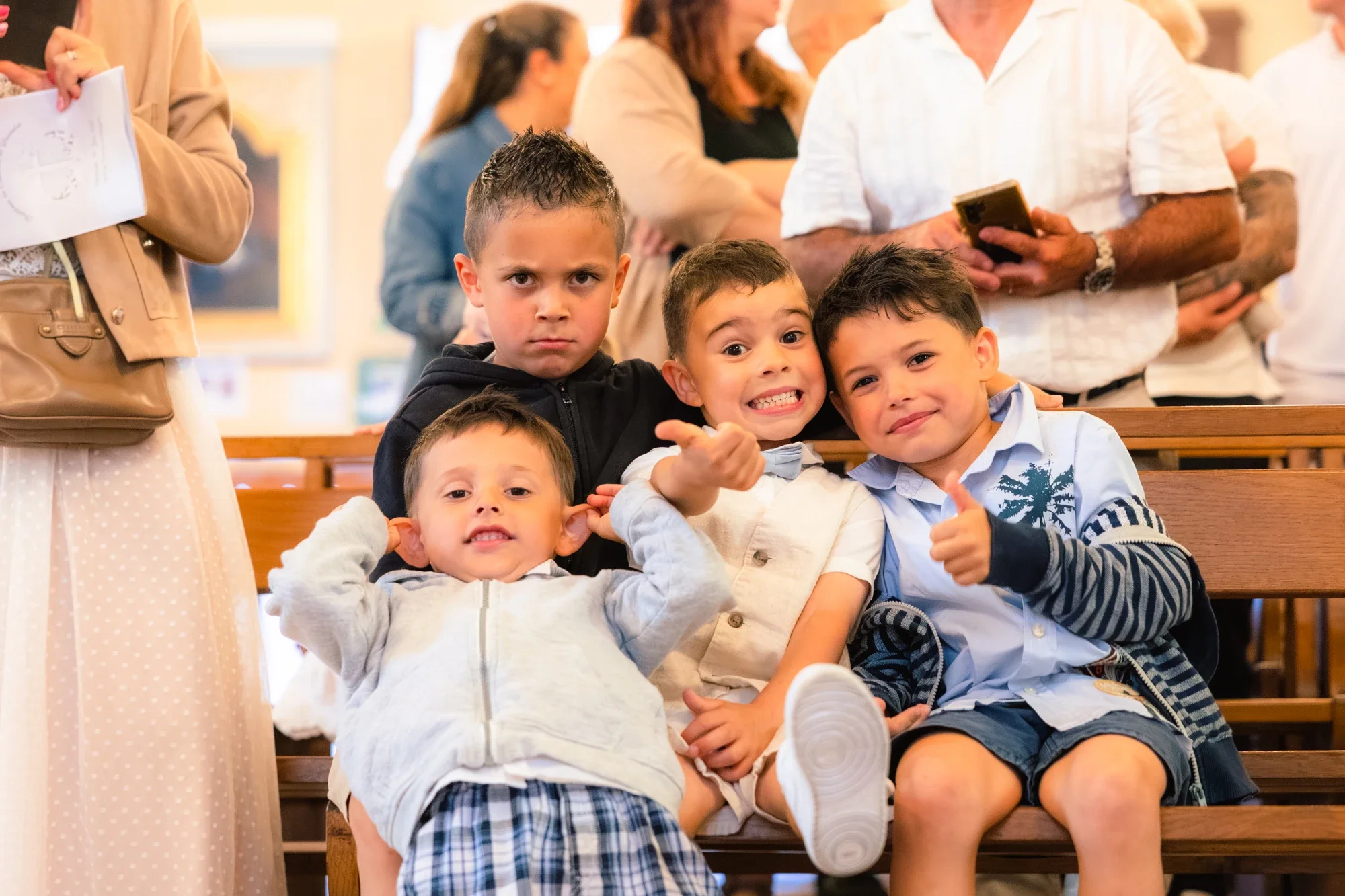 Groupe d'enfants assis sur des bancs, souriants et posant pour la photo dans un environnement intérieur, avec des adultes en arrière-plan.