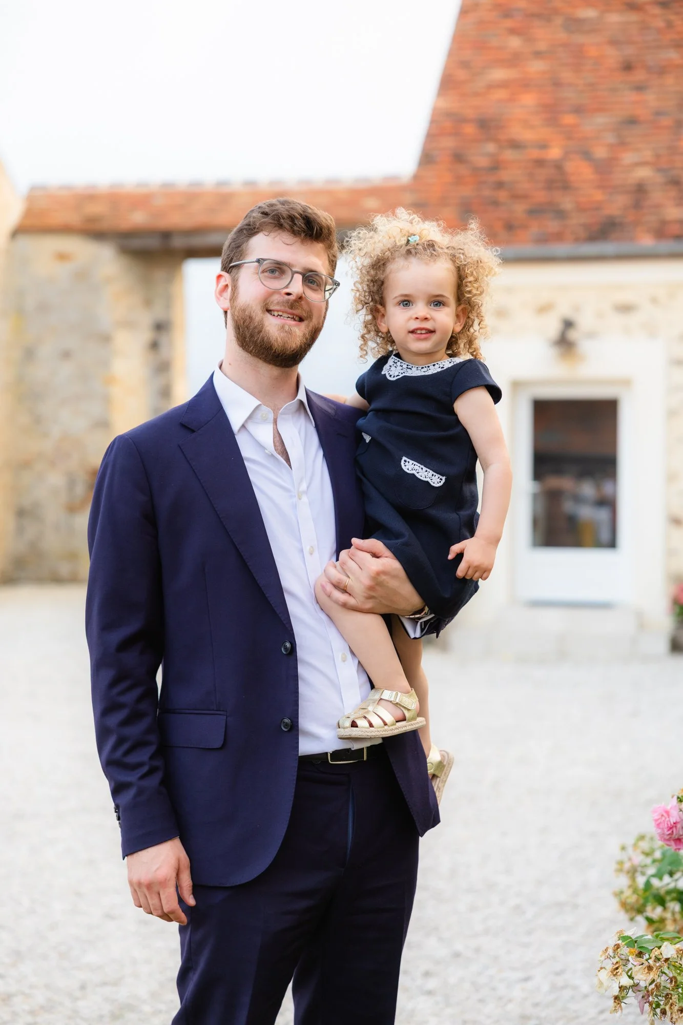Un homme en costume bleu tenant une petite fille aux cheveux bouclés rousse dans un centre-ville avec des bâtiments en pierre, une maison avec un toit en tuiles en arrière-plan, et des fleurs à droite.