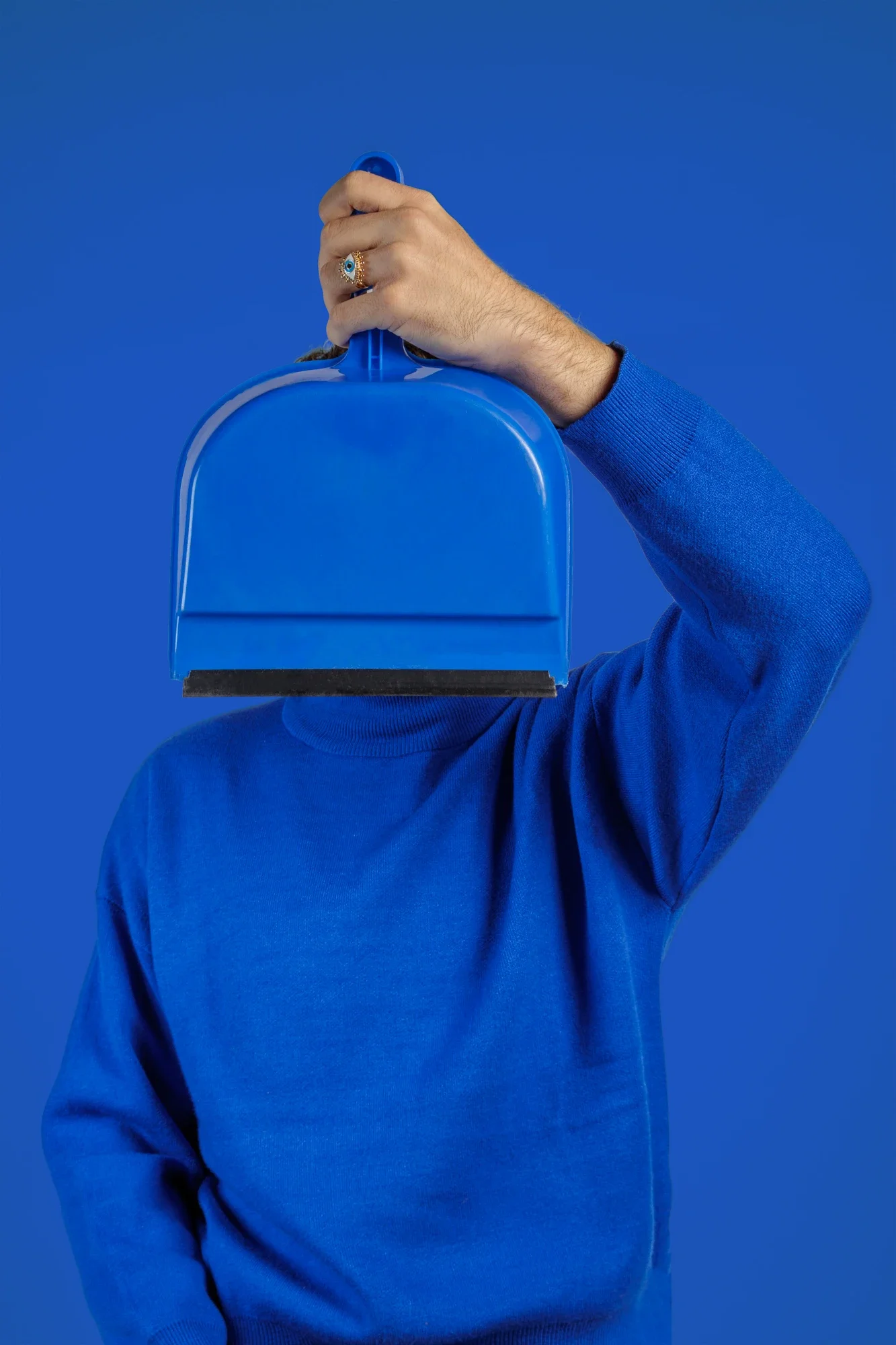 Personne en pull bleu tenant une pelle à poussière devant le visage, fond bleu.