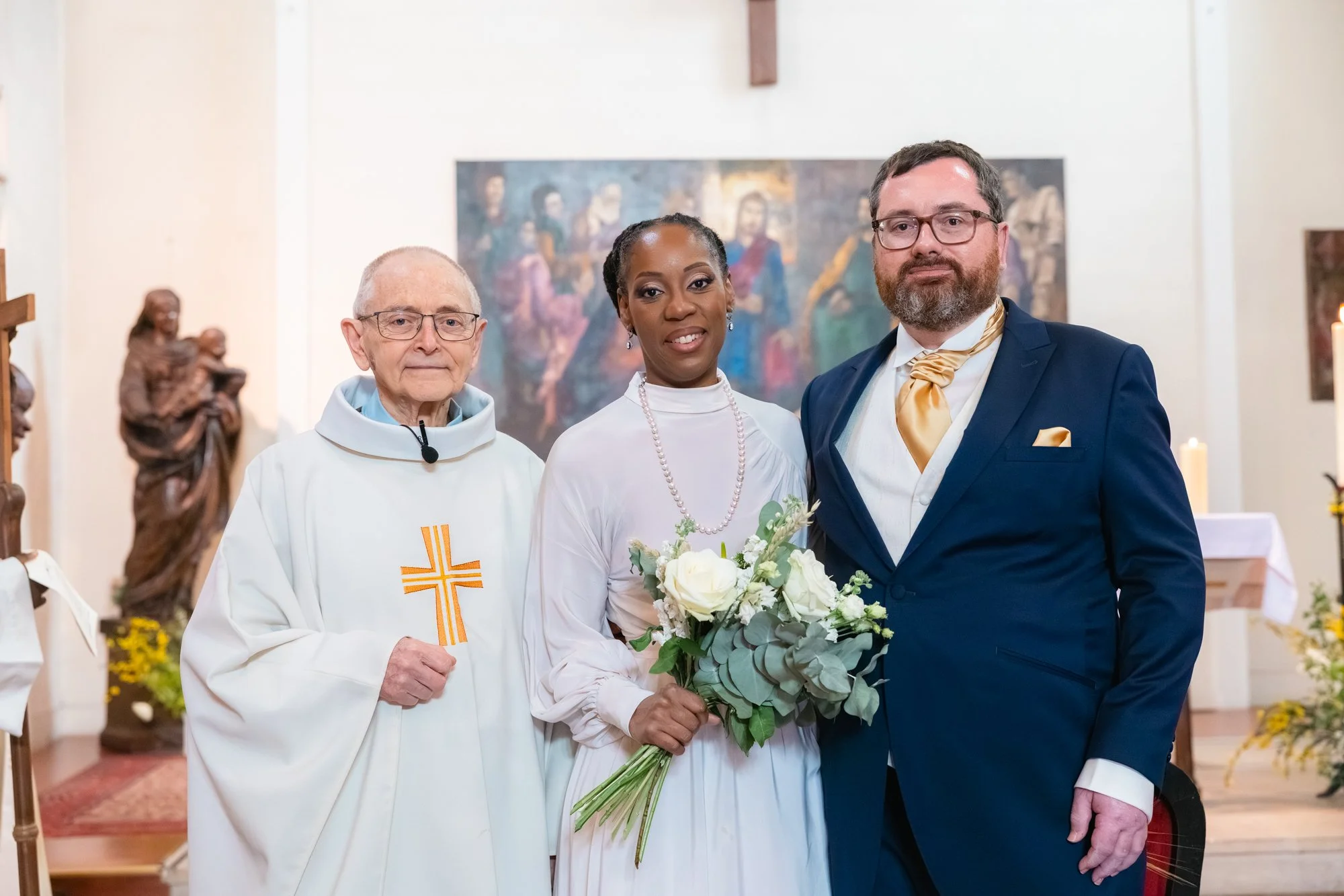 Trois personnes posent à l'intérieur d'une église. Une femme habillée en blanche, tenant un bouquet de fleurs, se tient entre un homme vêtu d'une costume bleu avec une écharpe dorée et un prêtre en tenue religieuse blanche avec une croix en or. En ar