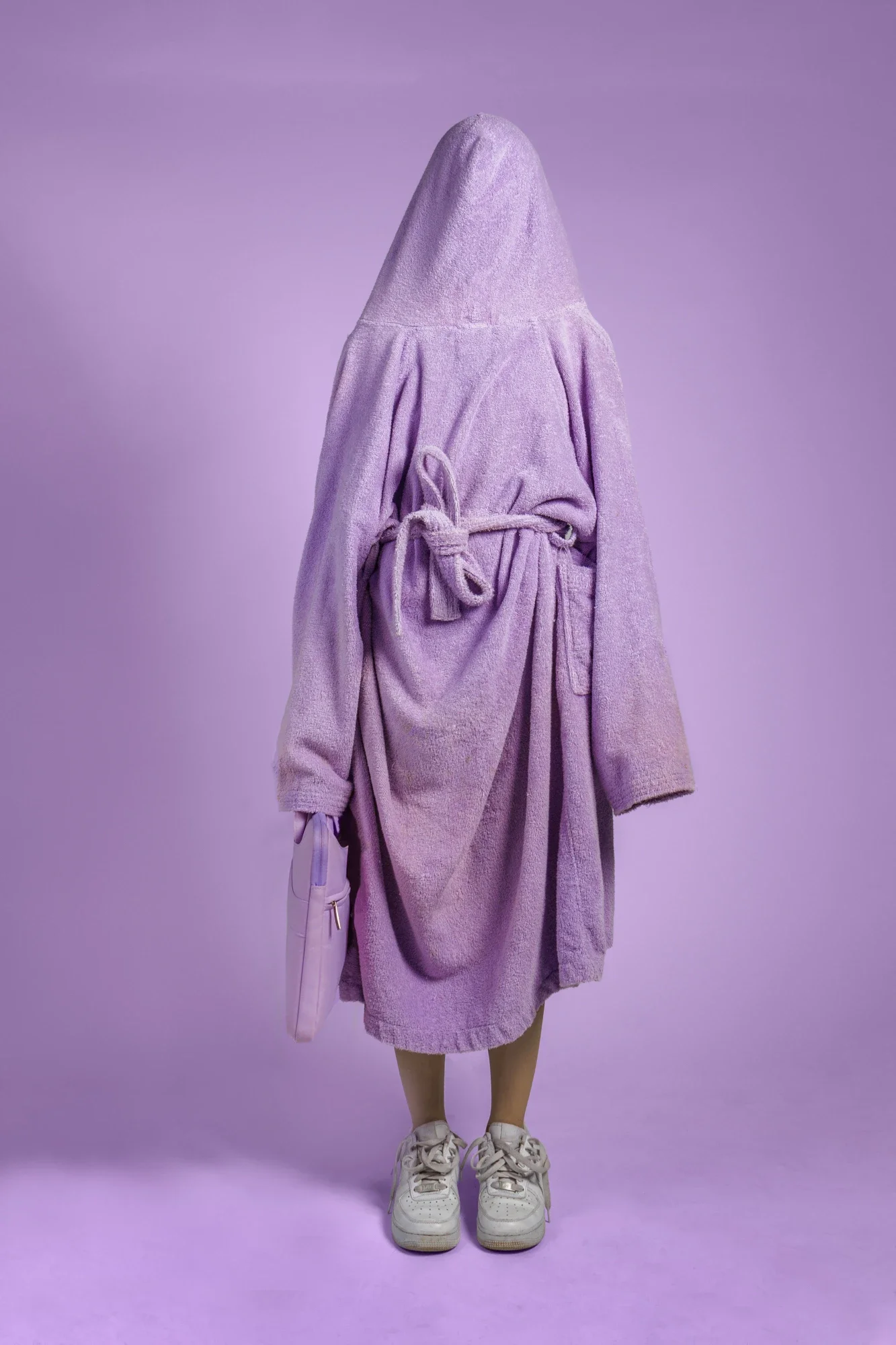 Mannequin portant un vêtement en peluche violet avec capuche, dans un fond violet. Mannequin portant des chaussures blanches, un sac à main violet et des pantalons beige.