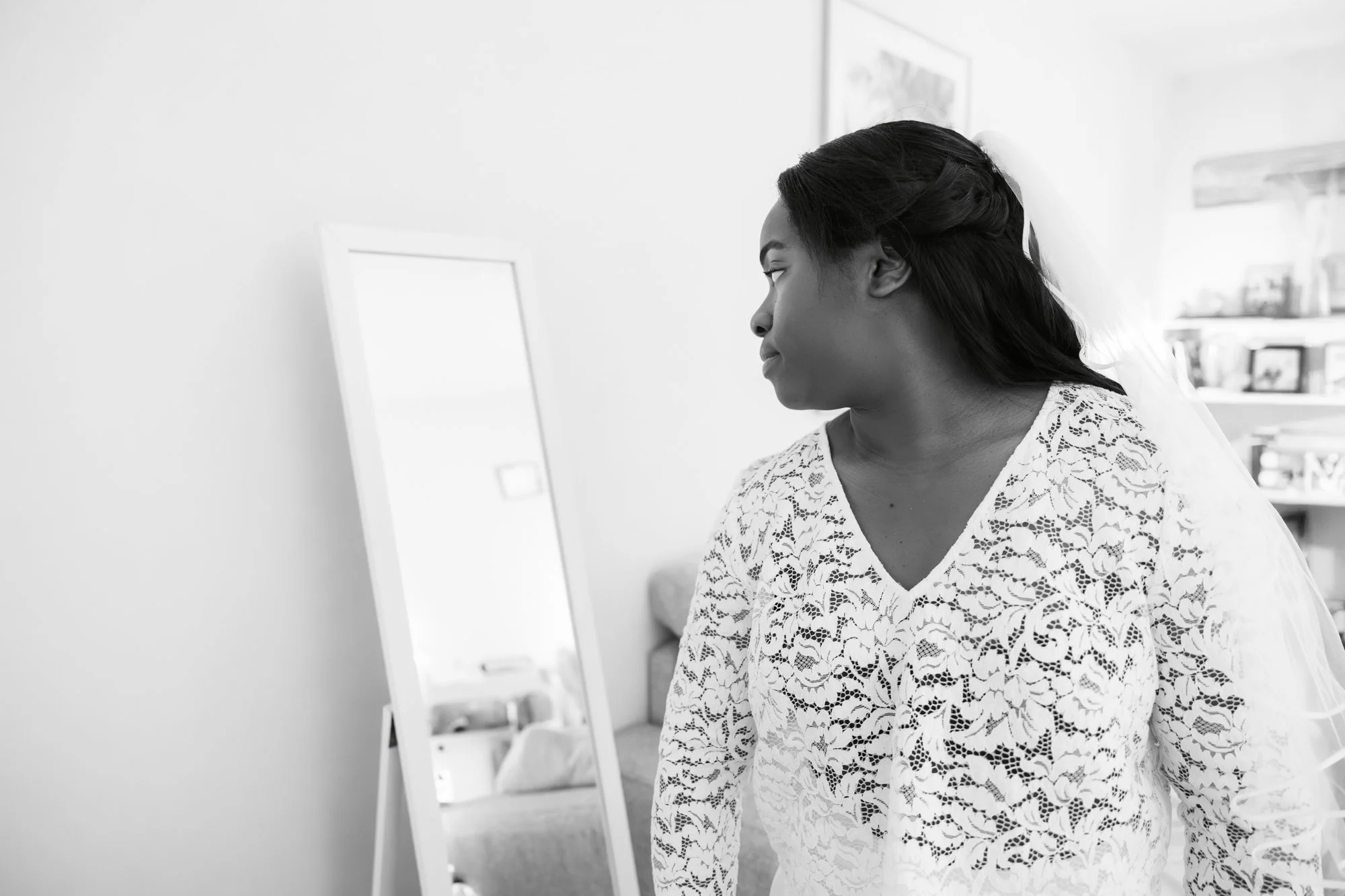 Une femme noire regarde dans un miroir, portant une robe en dentelle blanche.