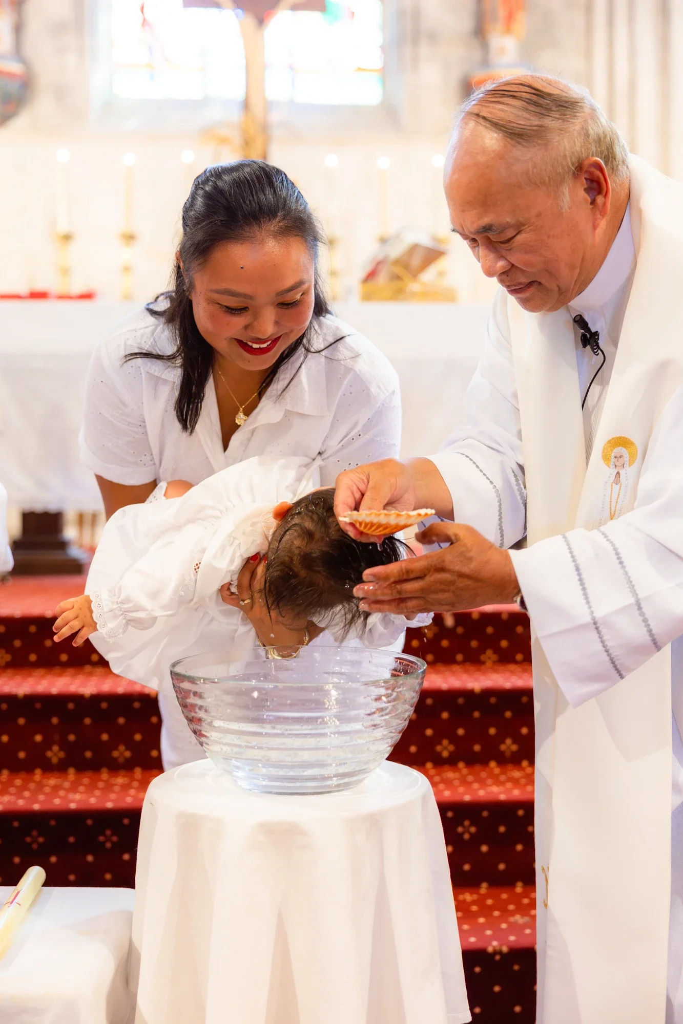 Un prêtre donne le baptême à un bébé dans un espace décoré avec une table où une bassine en verre est placée. La scène se déroule dans une église, avec des vitraux en arrière-plan.