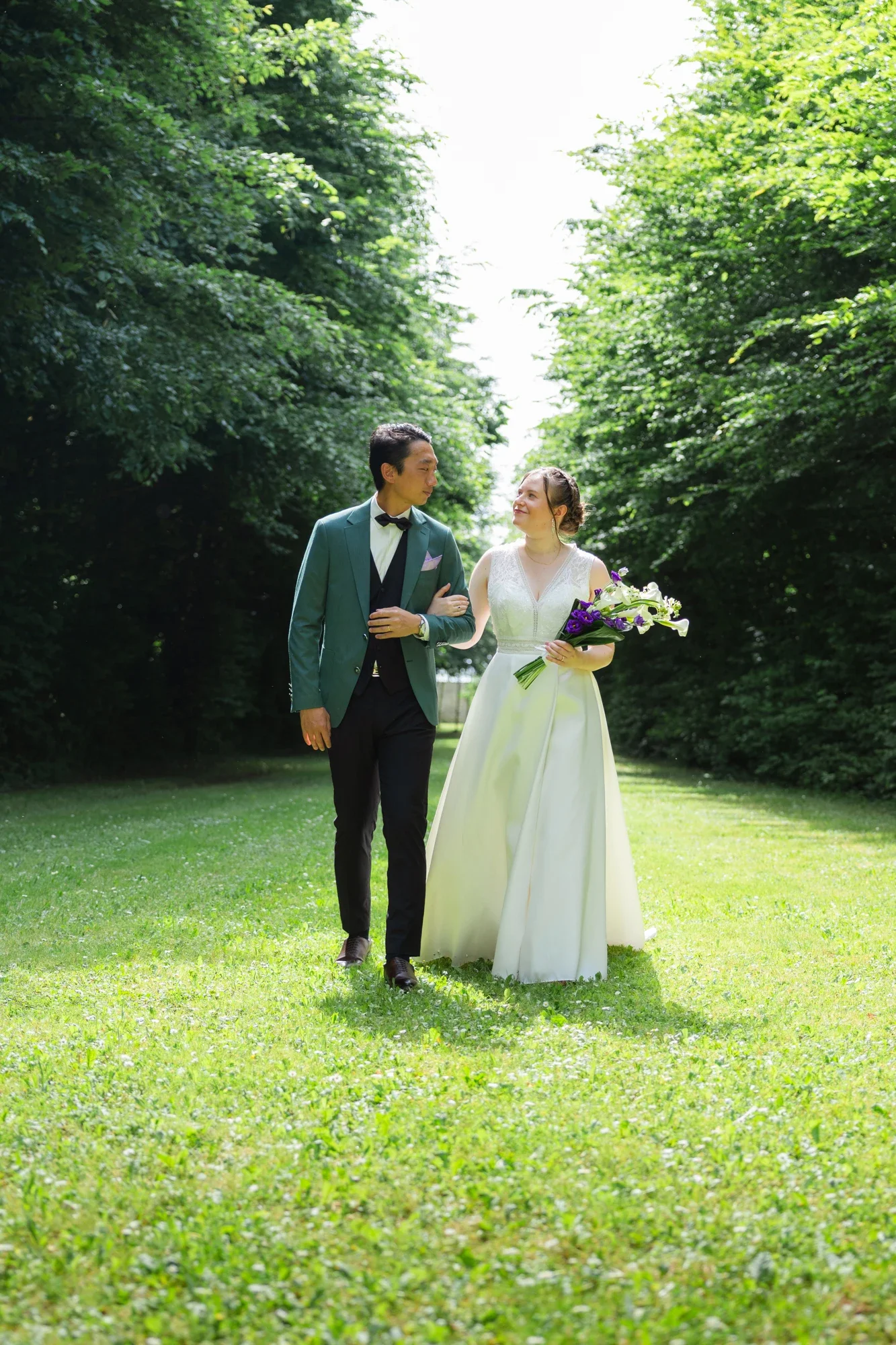 Un couple en vêtements de mariage marche dans un parc verdoyant. La femme porte une robe blanche et tient un bouquet de fleurs, l'homme porte un costume vert avec une chemise blanche et un nœud papillon.