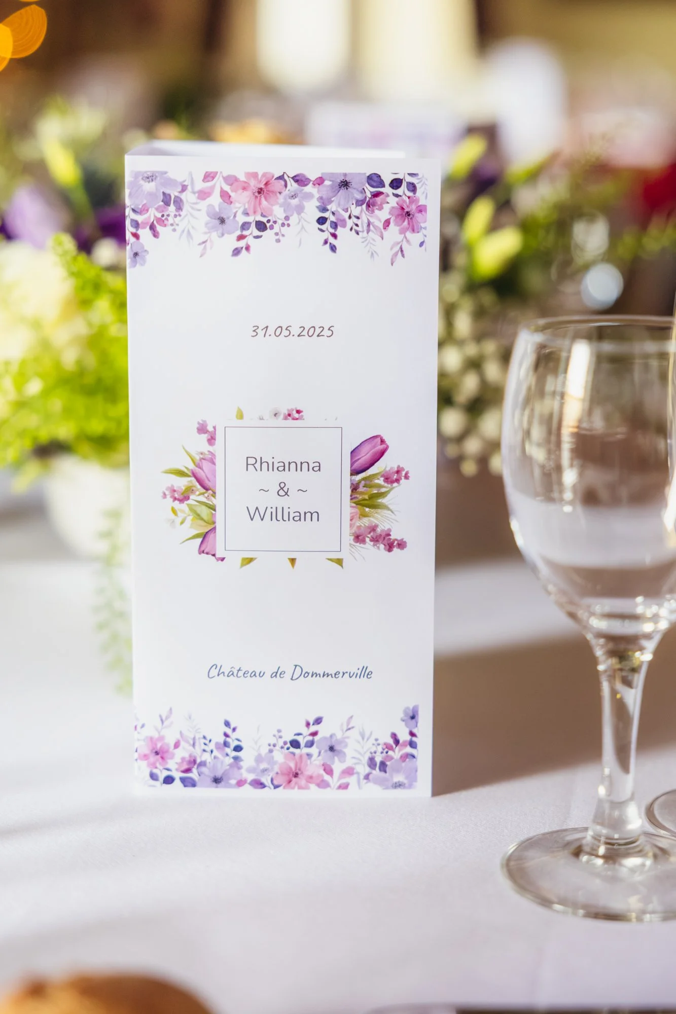Carte de mariage avec des fleurs violettes et roses, les noms Rhianna & William, la date 31.05.2025, sur une table avec un verre d'eau, et un bouquet de fleurs en arrière-plan.