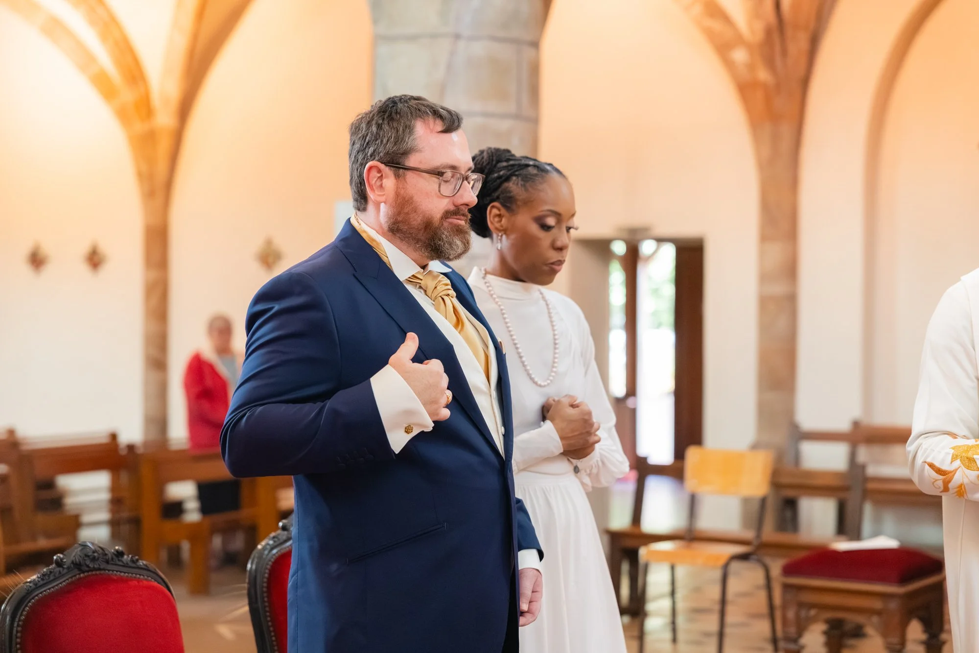 Deux personnes prient dans une église, l'homme en costume bleu et la femme en robe blanche, avec une ambiance chaleureuse et des bancs en bois velvet dans le fond.