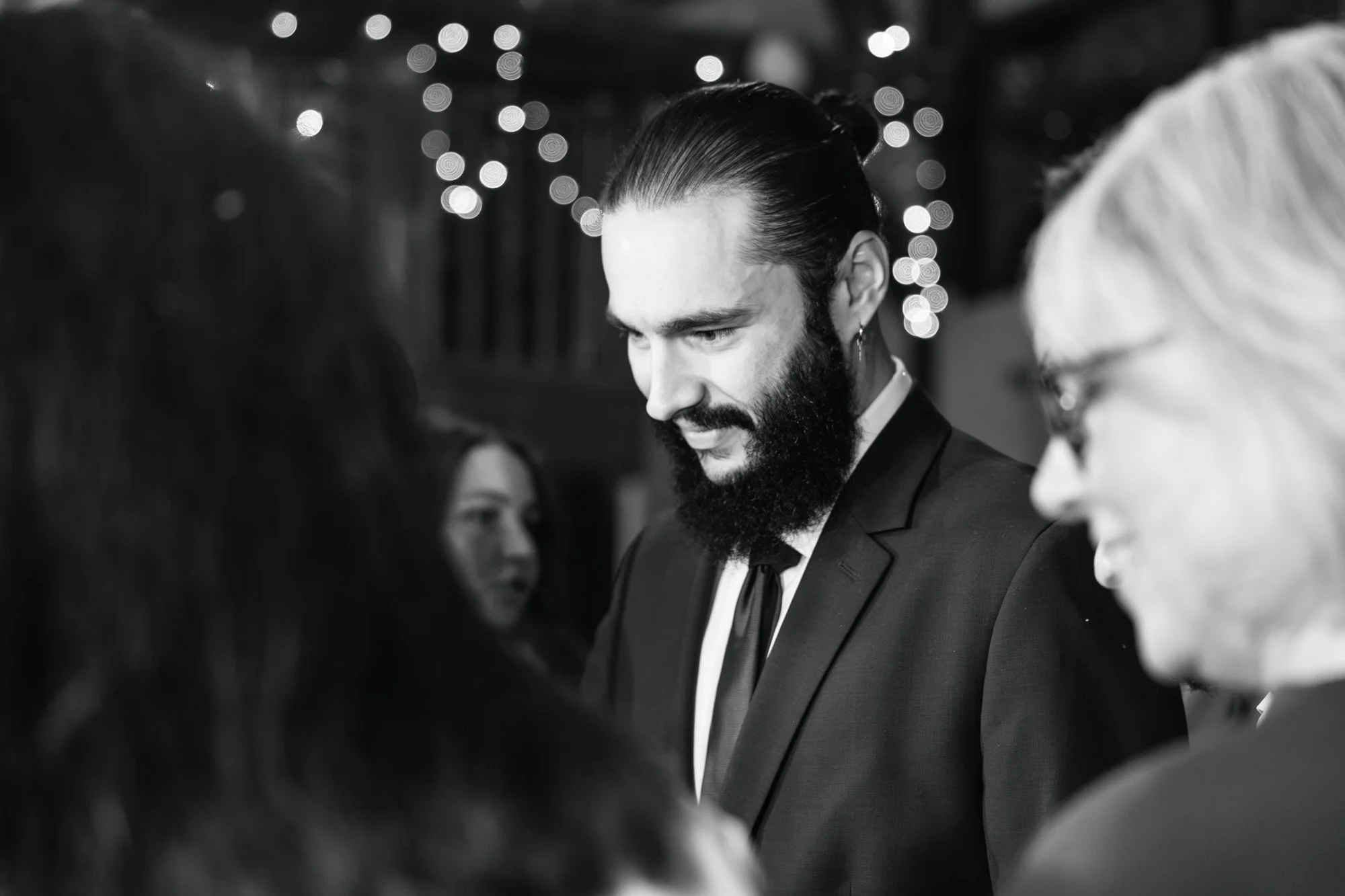 Un homme avec une barbe coiffée en arrière, portant un costume noir et une cravate, souriant lors d'une occasion festive avec d'autres personnes environnantes. En arrière-plan, des lumières décoratives floues.