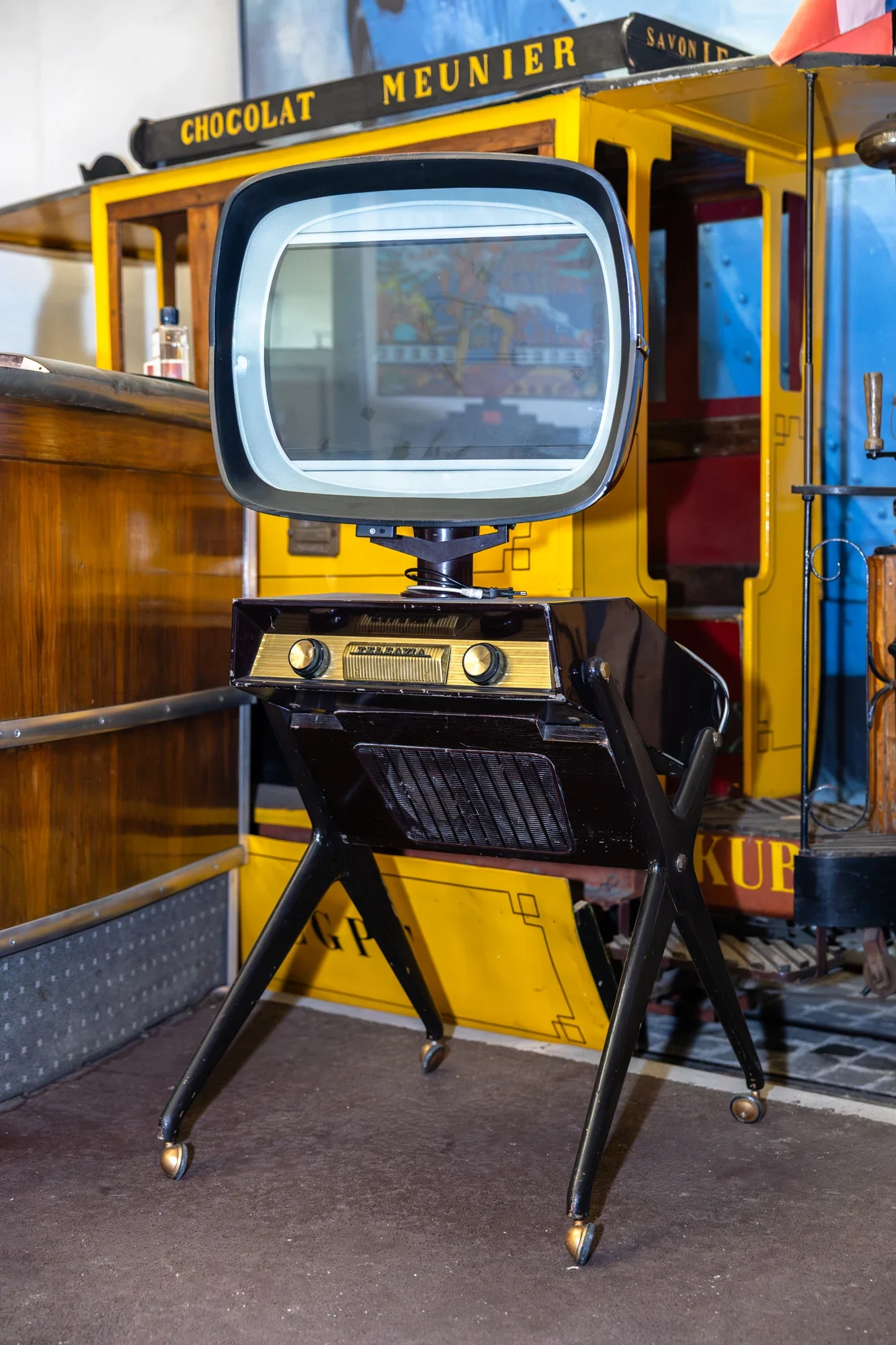 Un ancien jeu de flipper avec un écran en plastique et une console en métal, placé sur une chaise en plastique noire, dans un espace avec un décor vintage, notamment un stand en bois et un panneau jaune avec des mots "Chocolat Meunier".