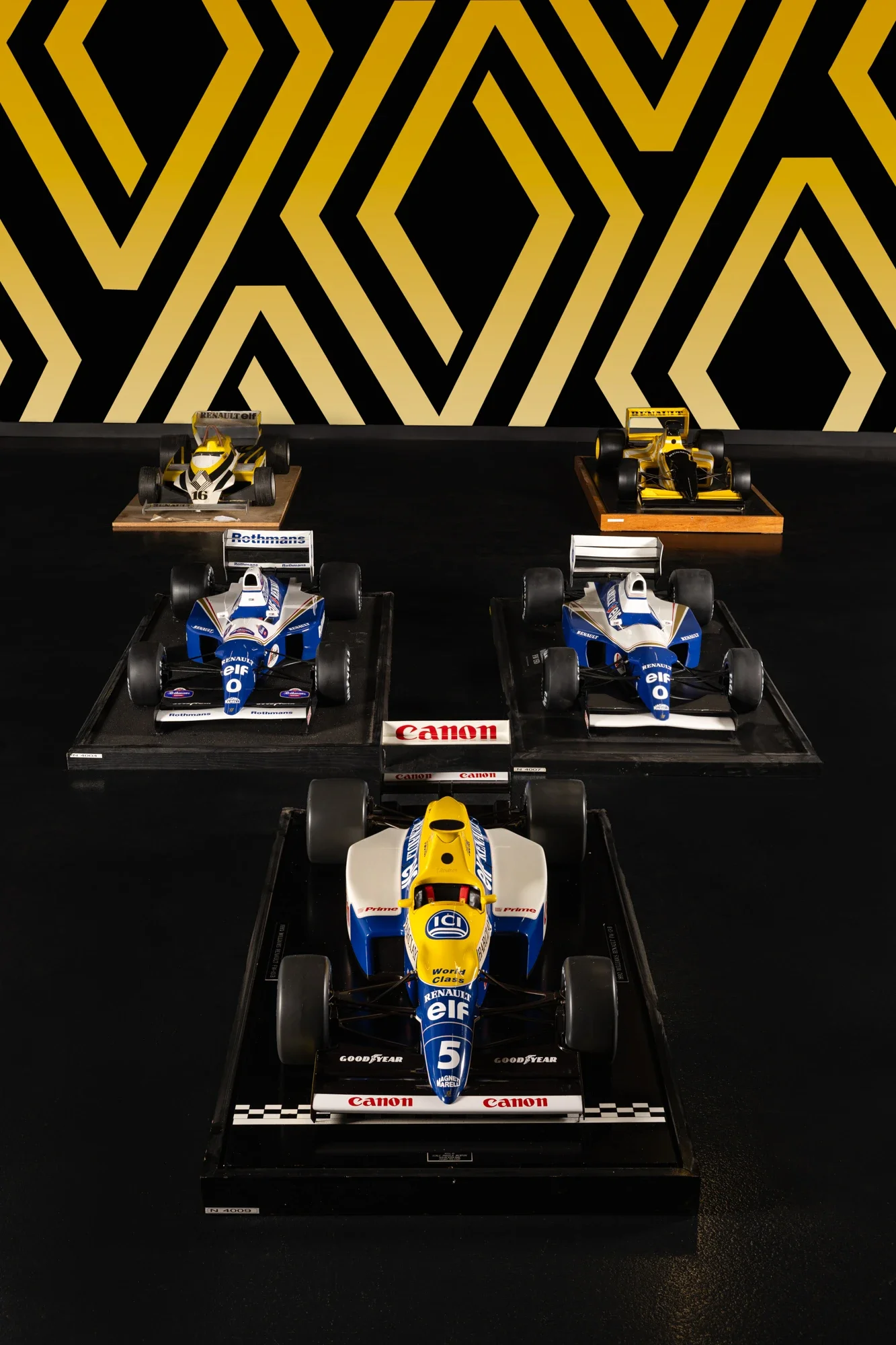 Une exposition de voitures de course classiques, principalement des Formules 1, exposées sur des plateformes, avec un mur à motif géométrique noir et or en arrière-plan.
