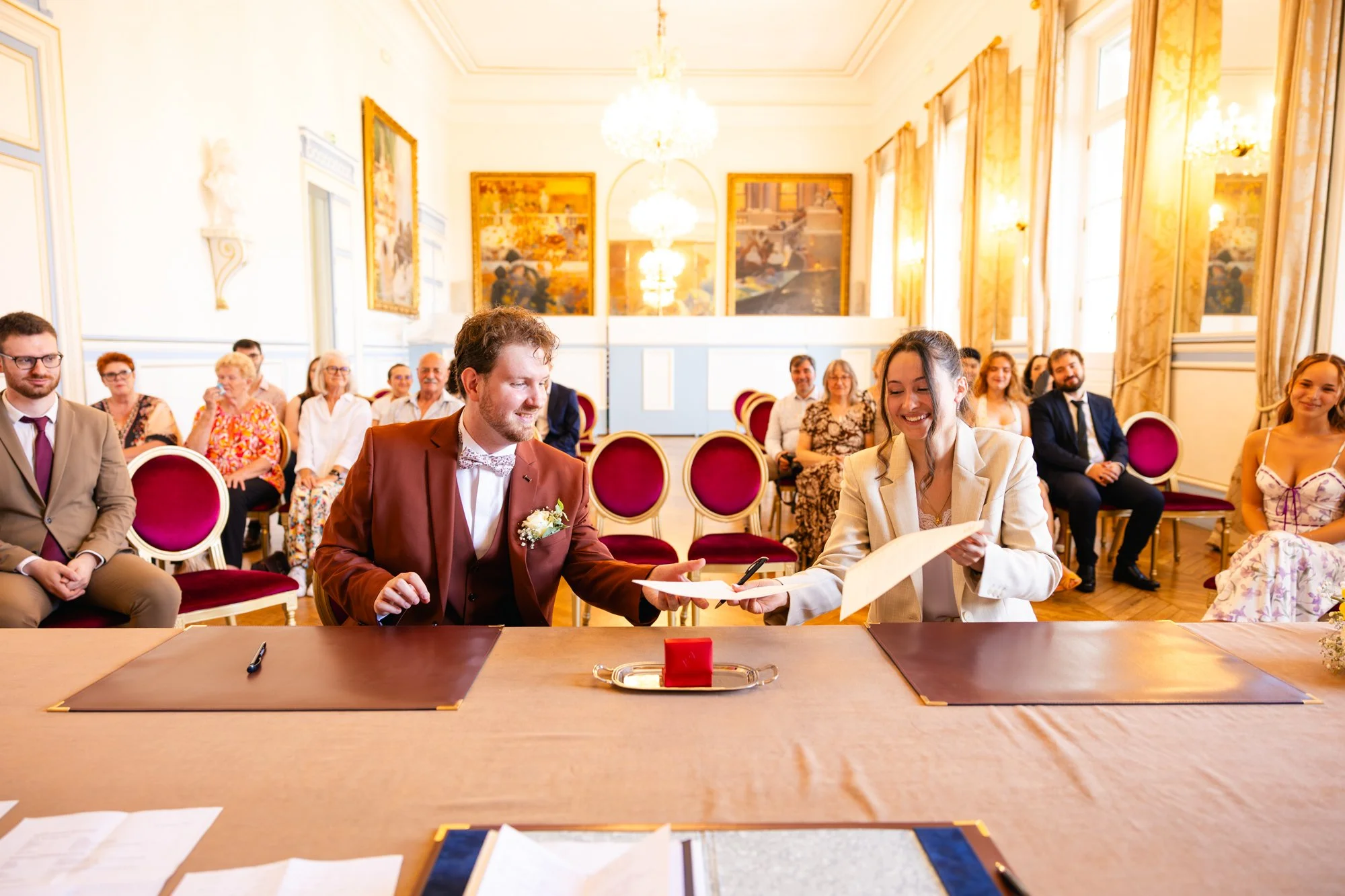 Un couple échange un document lors d'une cérémonie de mariage dans une salle élégante, entourés par des invités assis.