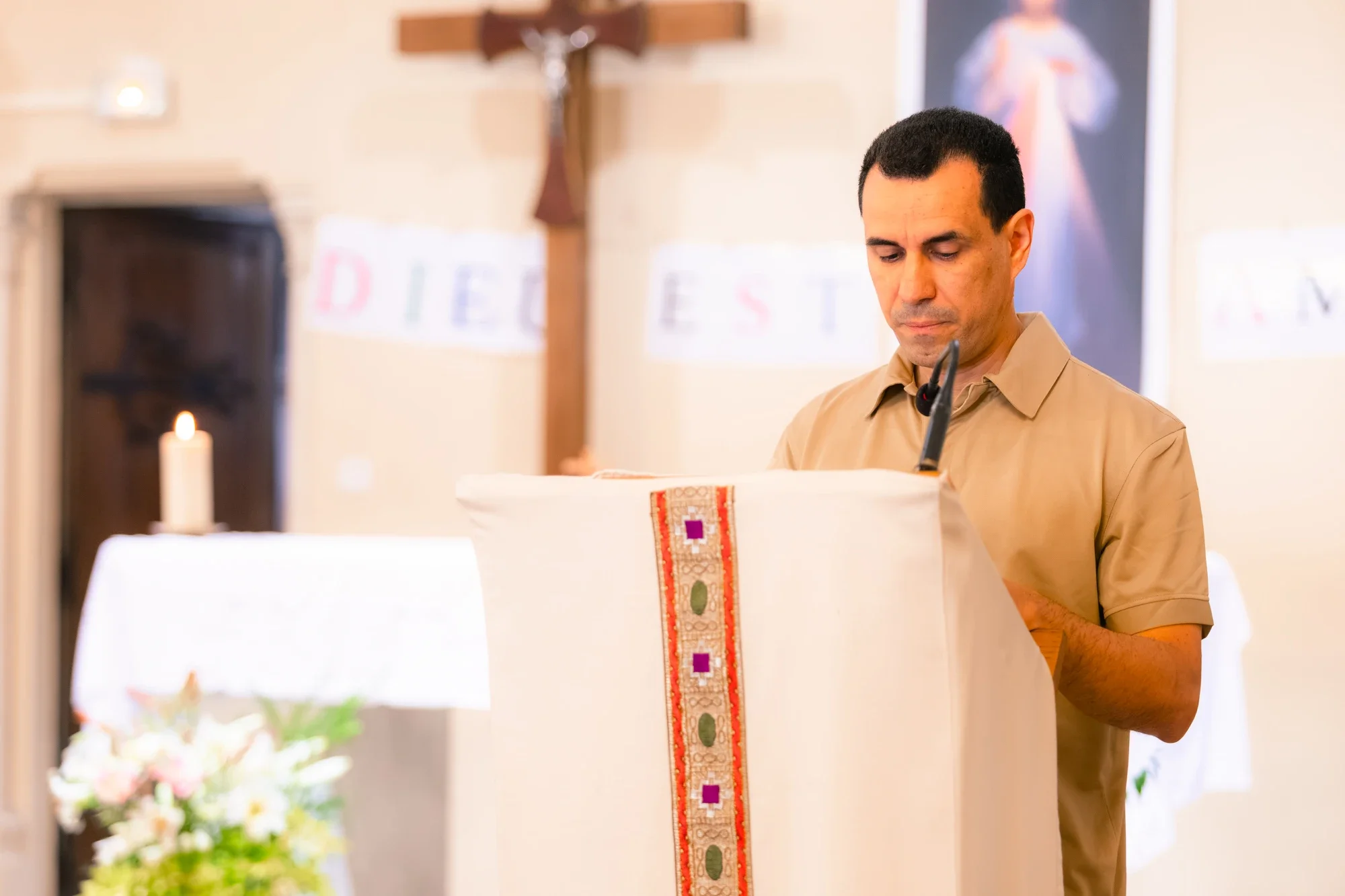 Un homme debout dans une église, probablement en train de prier ou de méditer, avec une croix en bois en arrière-plan, une bougie allumée, et des fleurs devant lui.
