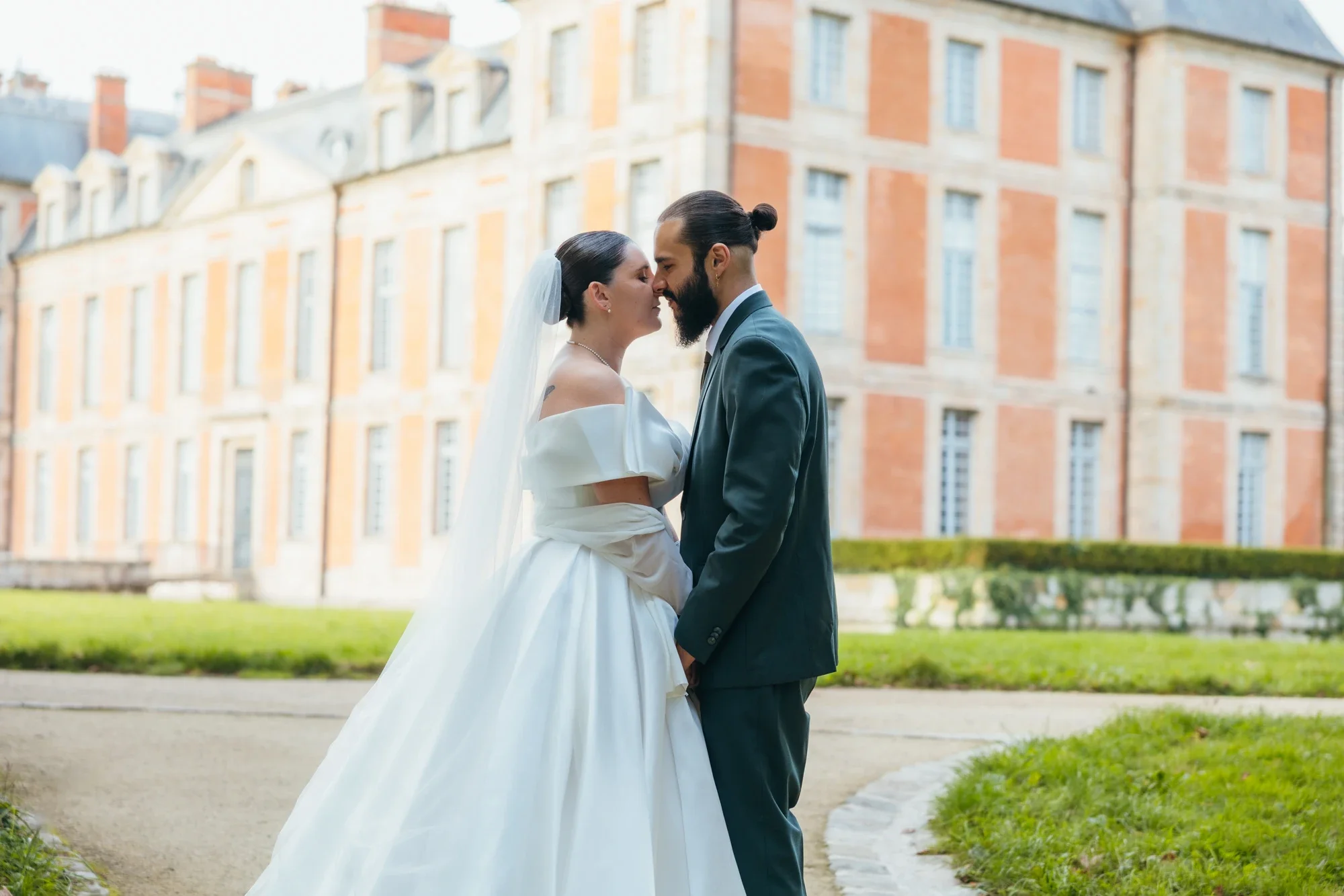 Un couple en mariage, la femme porte une robe blanche et un voile, et l'homme porte un costume gris, ils se tiennent face à face près d'un bâtiment historique en pierre rouge, lors d'une séance photo.