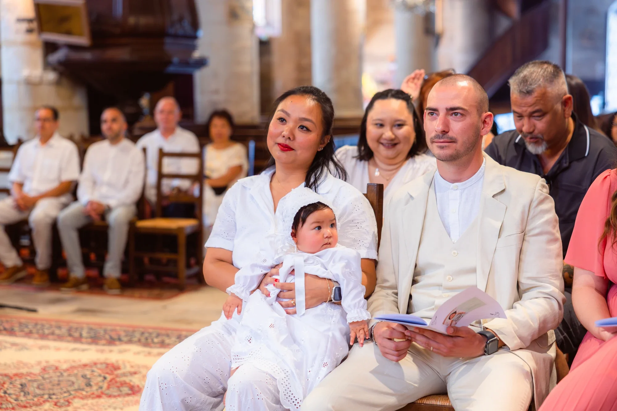 Groupe de personnes assises dans une salle, en attente d'une cérémonie, avec au premier plan une femme tenant un bébé en robe blanche et un homme en habit clair, tous deux concentrés, dans une ambiance religieuse ou cérémonielle.