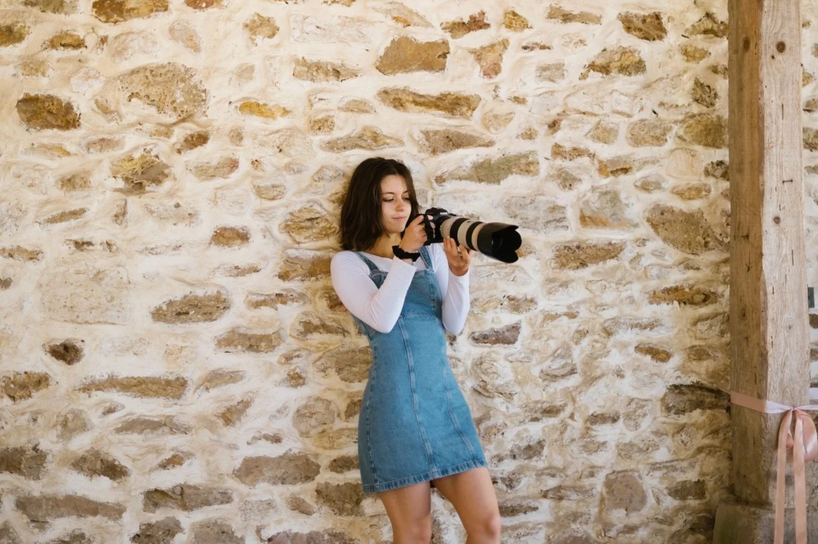 Jeune femme avec un appareil photo à objectif long, debout contre un mur en pierre dans un intérieur rustique.