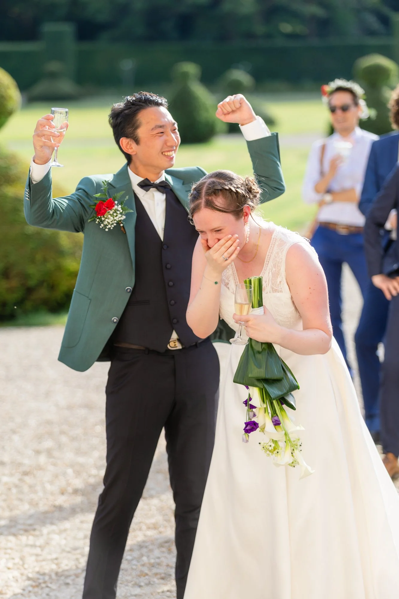 Un couple de mariés lors d'une cérémonie en plein air, la femme rit et couvre sa bouche avec sa main, tenant un bouquet de fleurs. L'homme porte un costume vert et un bouton de boutonnière rouge, il lève un verre. D'autres personnes en arrière-plan s