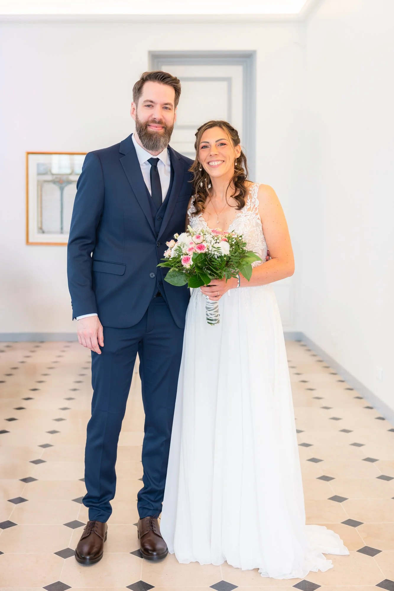 Un couple lors de leur mariage, debout dans une salle lumineuse. La femme porte une robe de mariée blanche et tient un bouquet de fleurs roses et blanches. L'homme porte un costume bleu foncé avec une cravate. Tous deux sourient.