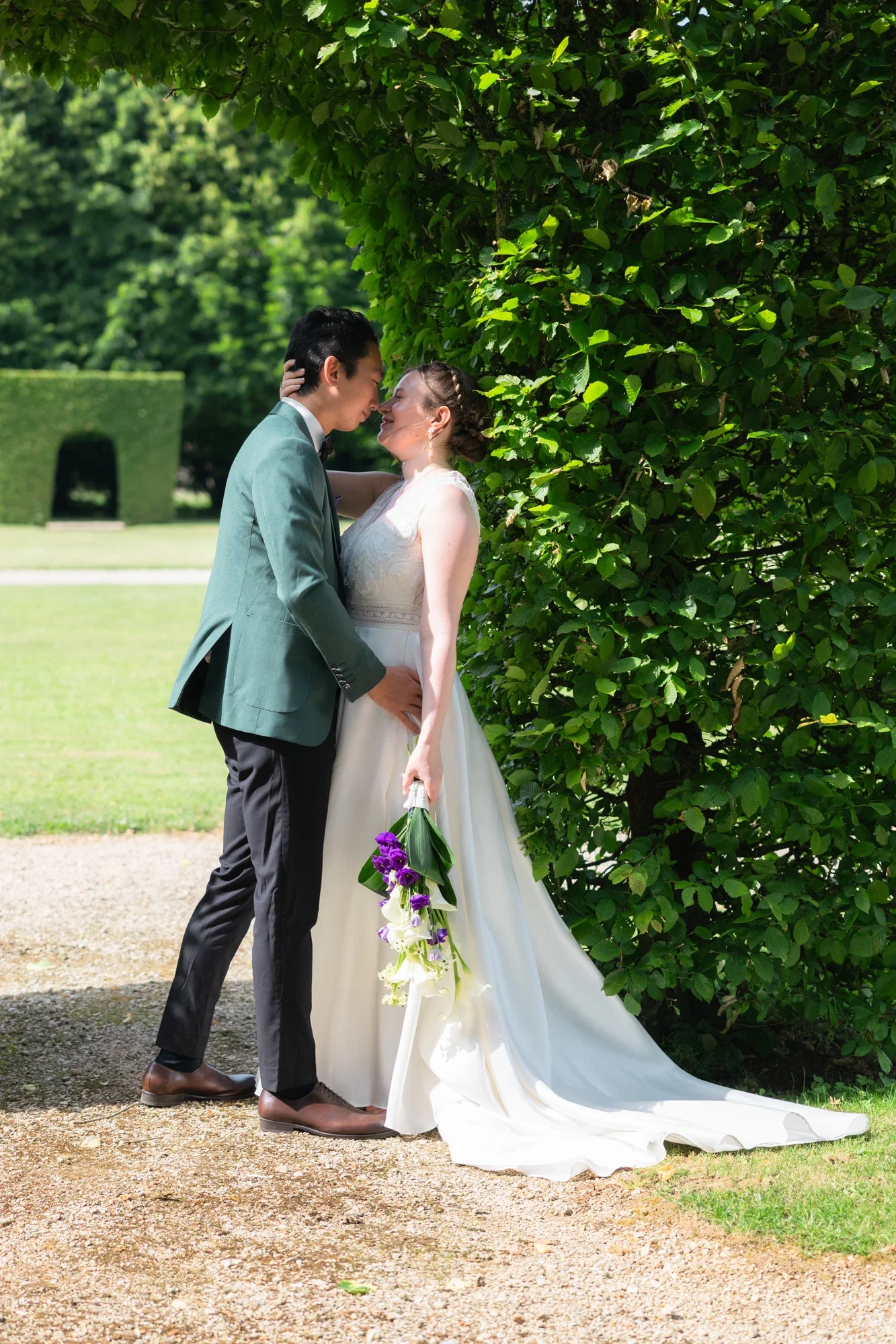 Un couple de mariés en train de s'embrasser devant un buisson vert dans un parc en plein air, la femme tenant un bouquet de fleurs blanches et violettes.