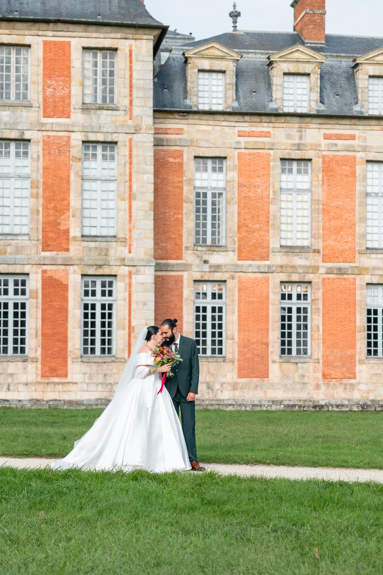 Un couple de mariés debout sur l'herbe devant un bâtiment en briques, la mariée en robe blanche longue et le marié en costume sombre. La mariée tient un bouquet de fleurs colorées.