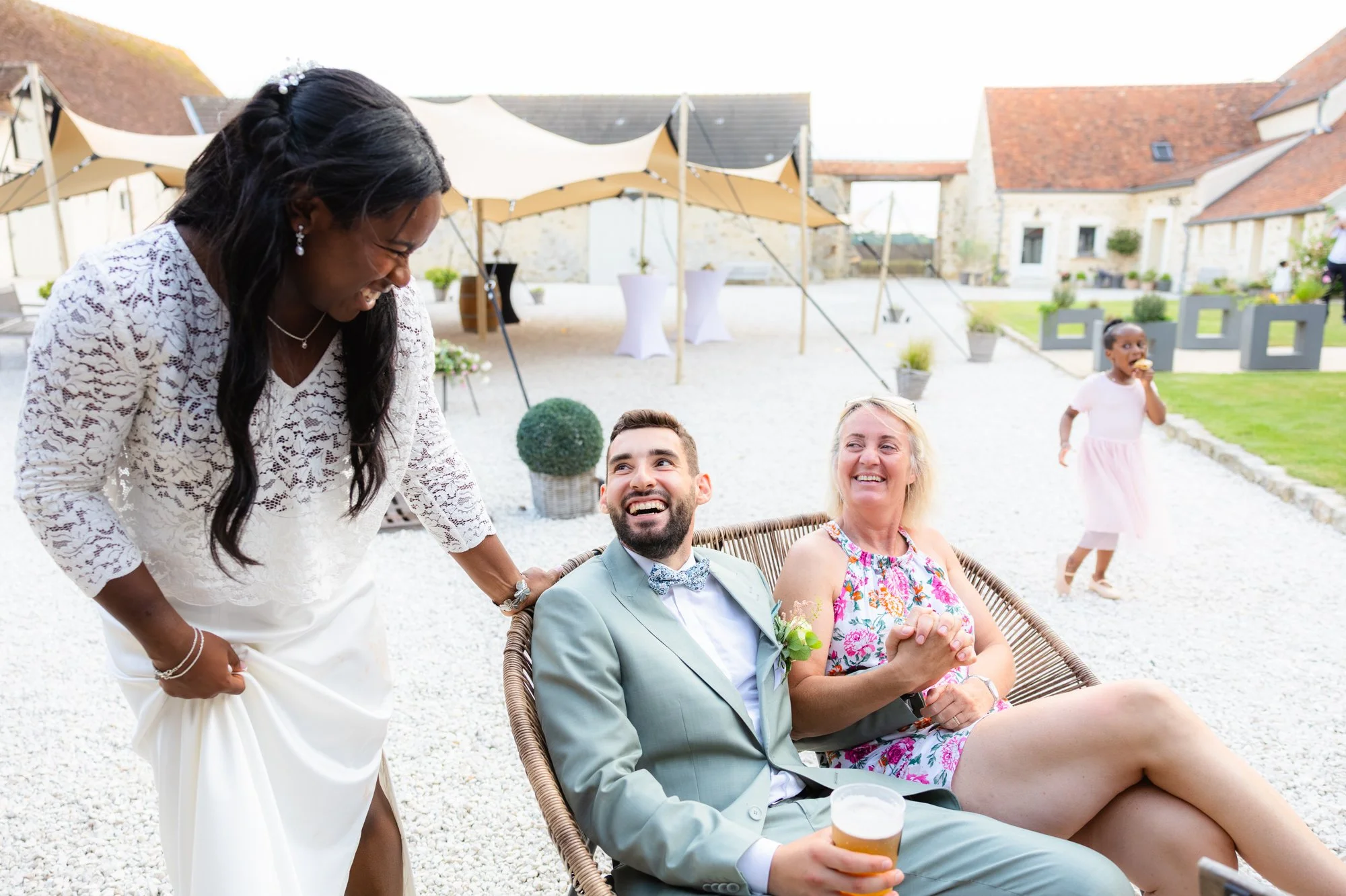 Un groupe de personnes à une fête en plein air, deux femmes et un homme assis, une femme avec une robe florale et l'homme avec un costume, tous souriants. Une autre femme en blanc les serre par la main, et une petite fille en robe rose mange un cupca