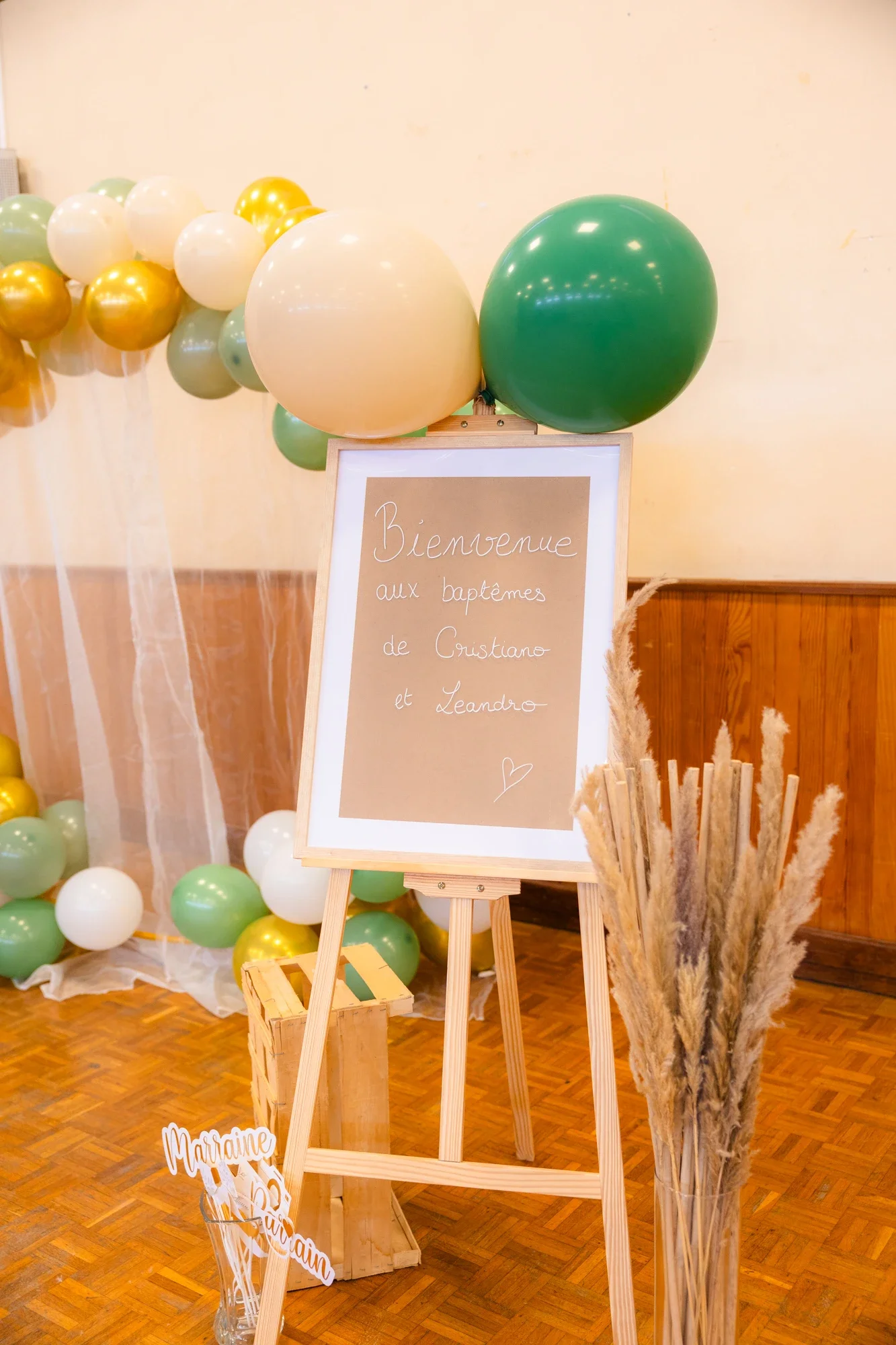 Décoration de baptême avec un panneau écrit 'Bienvenue aux baptêmes de Cristiano et Leandro' entouré de ballons verts, blancs, et dorés, et d'une vase avec des pampas
