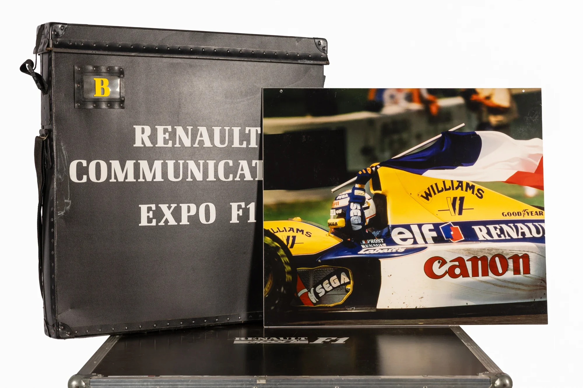 Une valise noire avec l'inscription "Renault Communication Expo F1" en blanc et un panneau publicitaire montrant une voiture de course Renault Williams avec des logos de sponsors comme Canon, Elf, et Michelin.