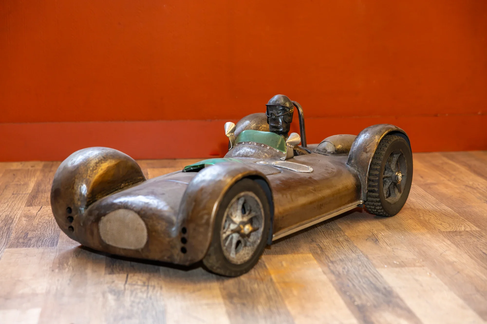 Voiture de course en métal vintage, avec un pilote en casque et lunettes, posée sur un sol en bois contre un mur orange.