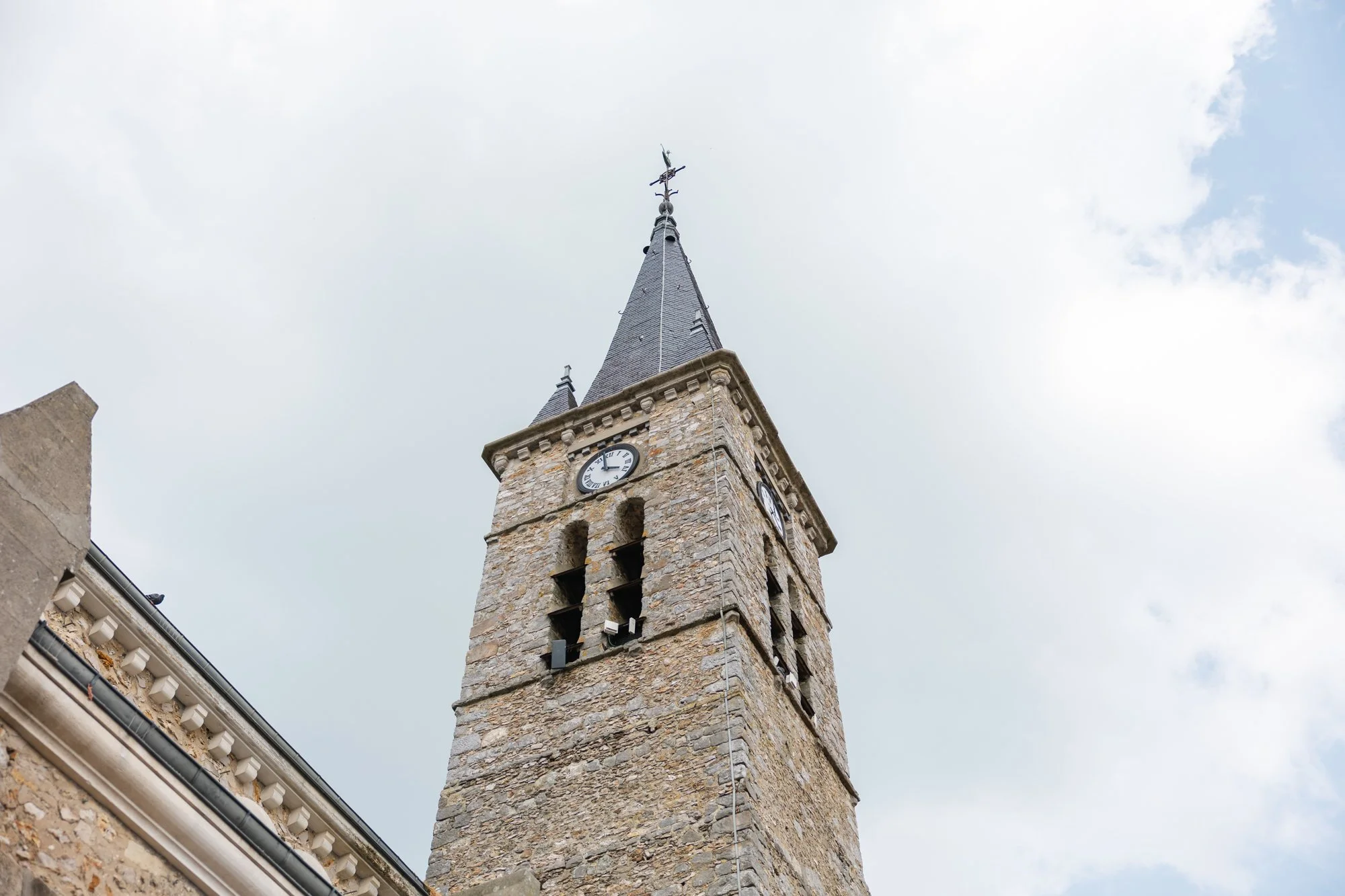 Une tour d'horloge en pierre avec un toit pointu en ardoise, surmontée d'une grande croix ou girouette, contre un ciel nuageux.