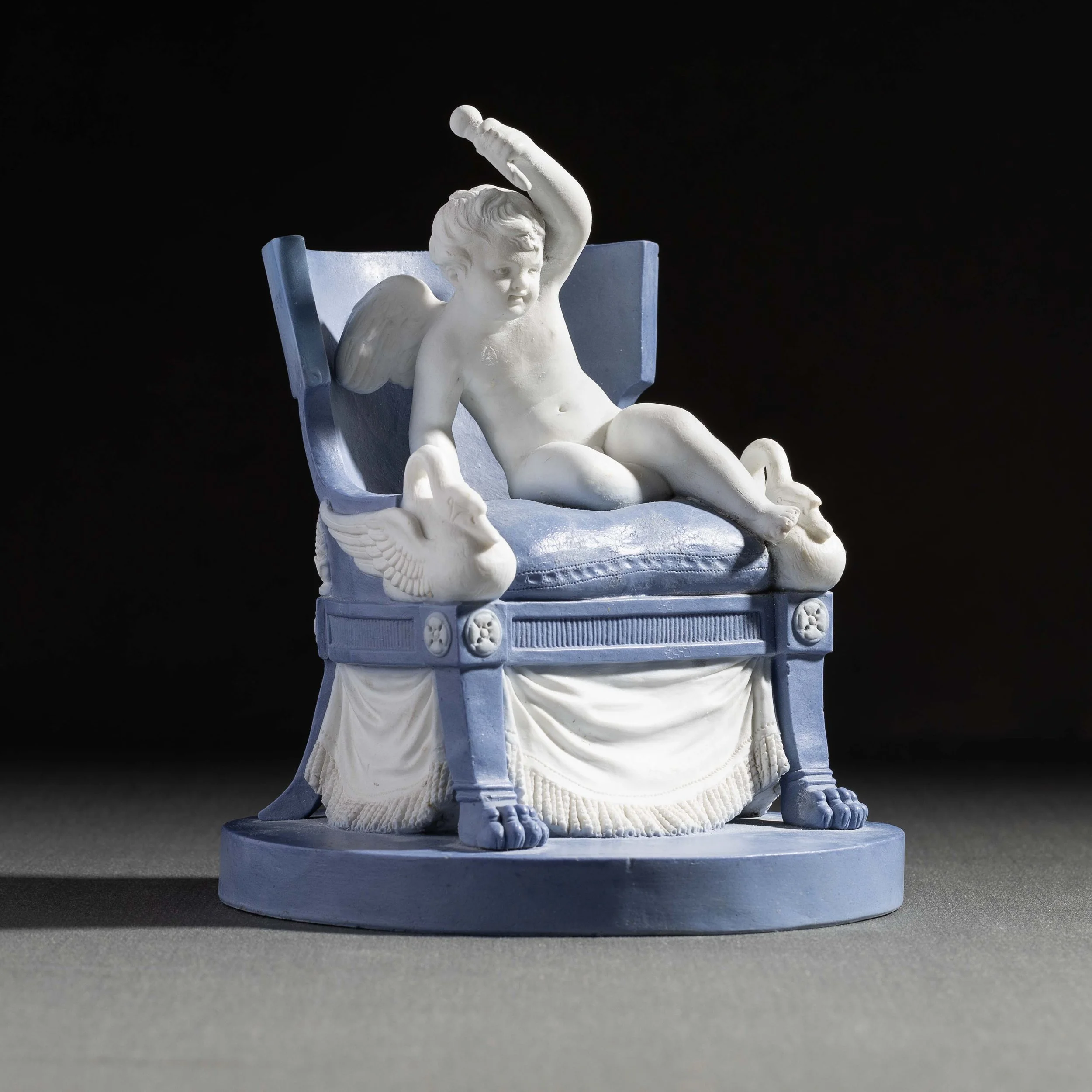 Statue blanche représentant un putto avec des ailes, assis sur un support en forme de coffre orné de motifs, contre un fond noir.