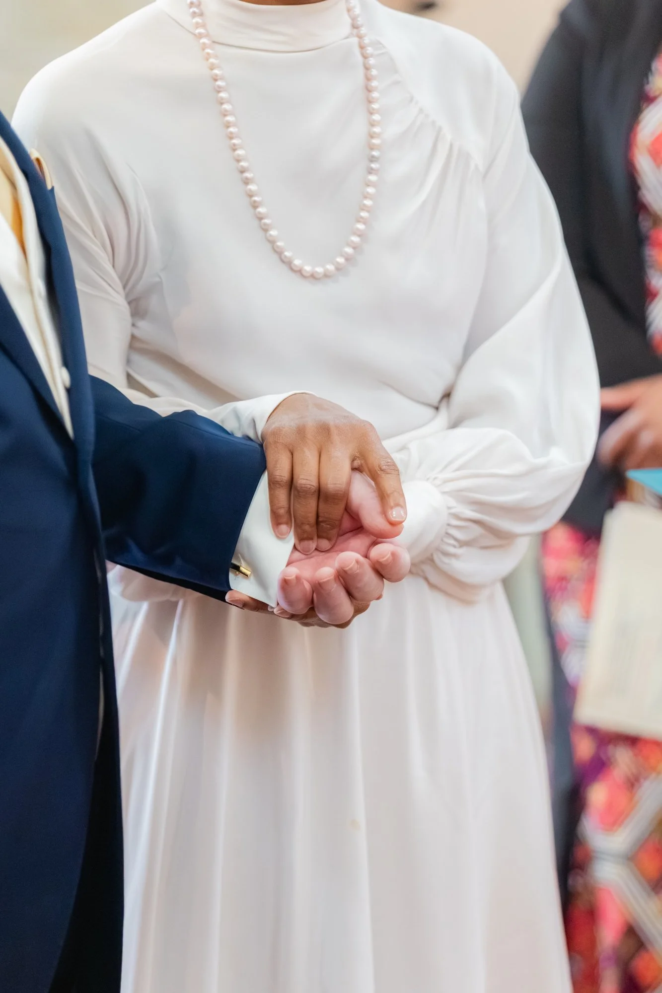 Groupe de personnes se tenant la main lors d'une réunion ou d'une cérémonie. La personne au centre porte une robe blanche avec un collier de perles roses, et d'autres vêtements sont visibles sur les côtés.