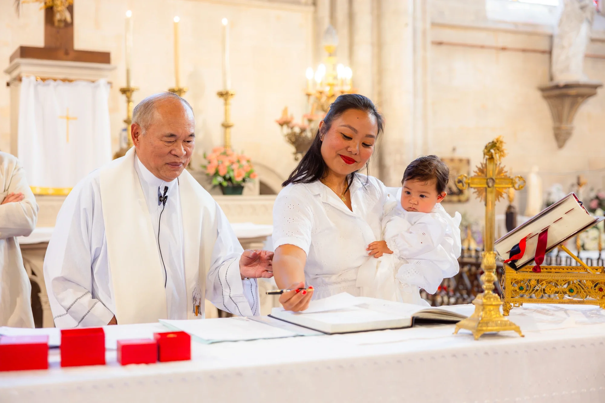 Une famille participe à une cérémonie religieuse dans une église. Un prêtre, une femme tenant un bébé, sont présents devant une table avec des objets de cérémonie, le tout dans un décor d'église avec des bougies et des fleurs.
