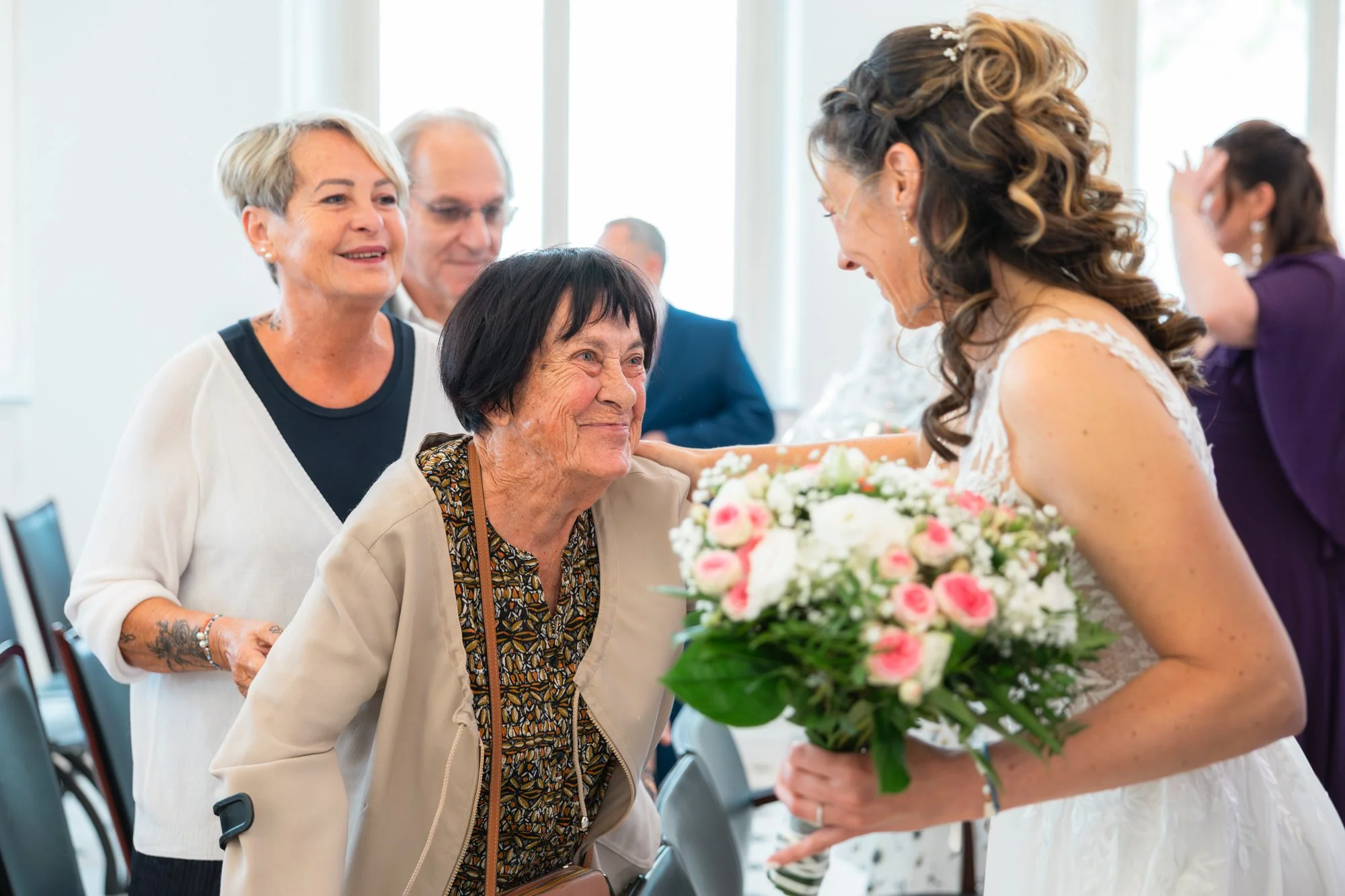 Une femme âgée souriante accepte un bouquet de fleurs offert par une mariée lors d'une cérémonie. Autour, d'autres personnes assistent à l'événement dans une salle lumineuse.