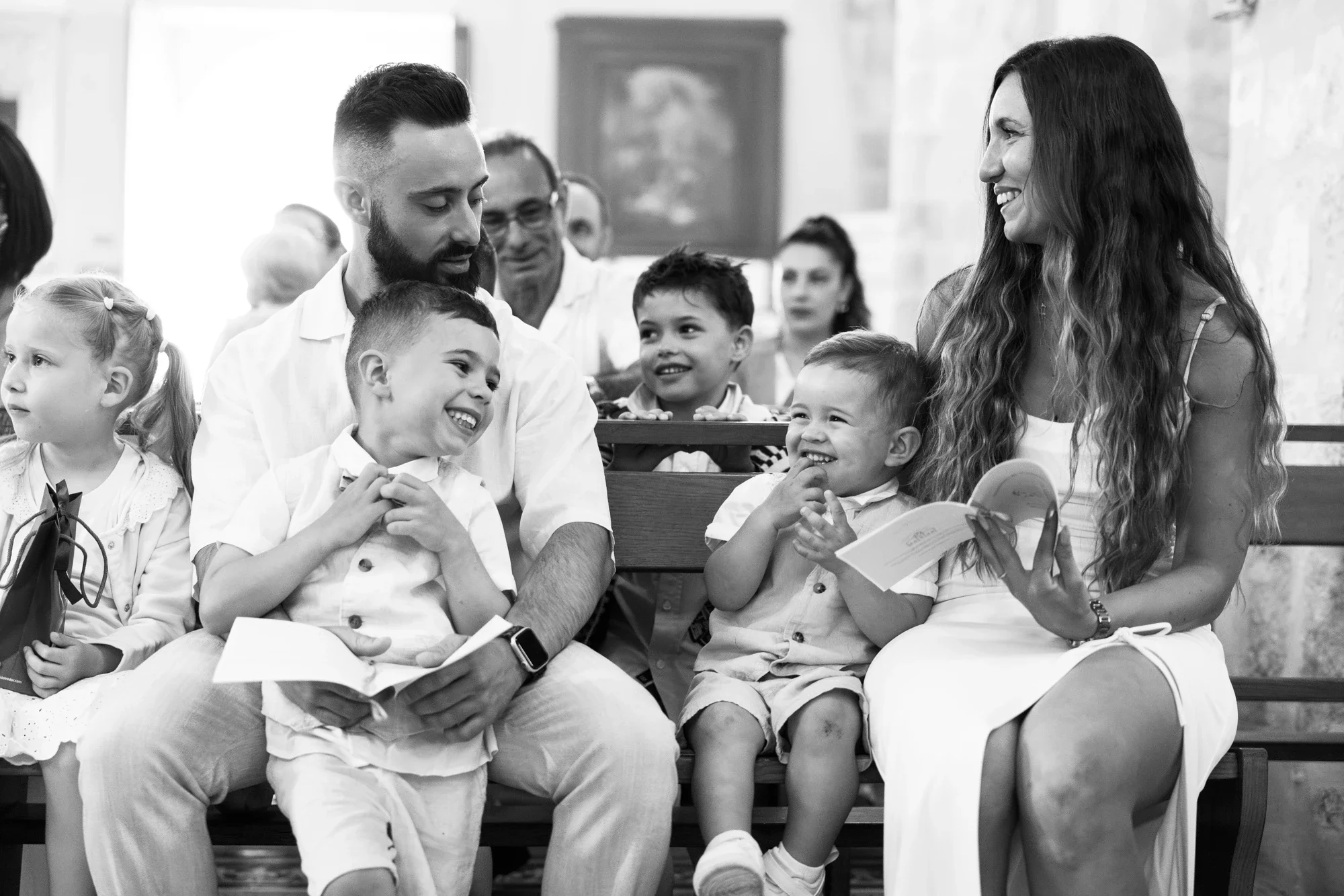 Une femme et un homme souriants assis dans une salle remplie d'enfants, la femme tenant un livre ouvert, tous semblent engagés dans une activité ou une conversation joyeuse.