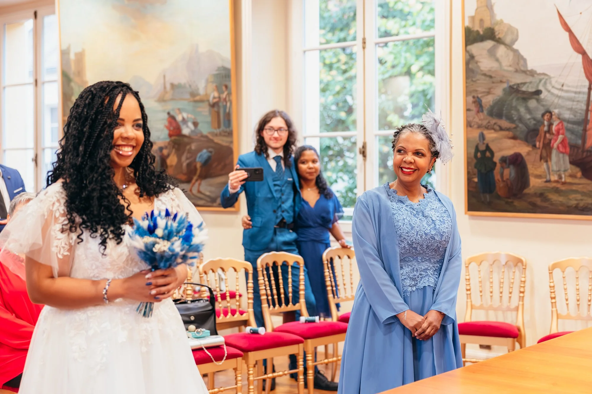 Cérémonie ou célébration avec des personnes souriantes, une femme en robe blanche avec des fleurs et une autre en bleu, dans une salle d'exposition avec des tableaux et des chaises en bois.