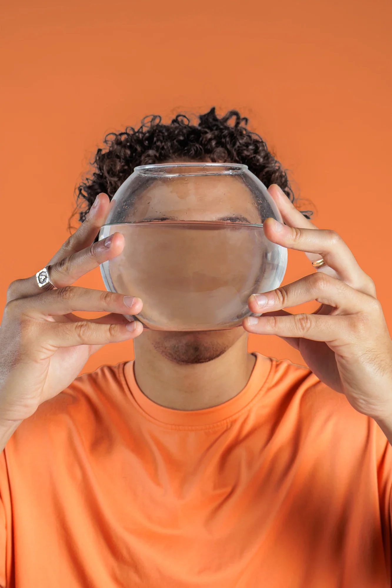 Jeune homme portant un t-shirt orange, tenant un verre en verre rond transparent rempli d'eau devant son visage, contre un fond orange.