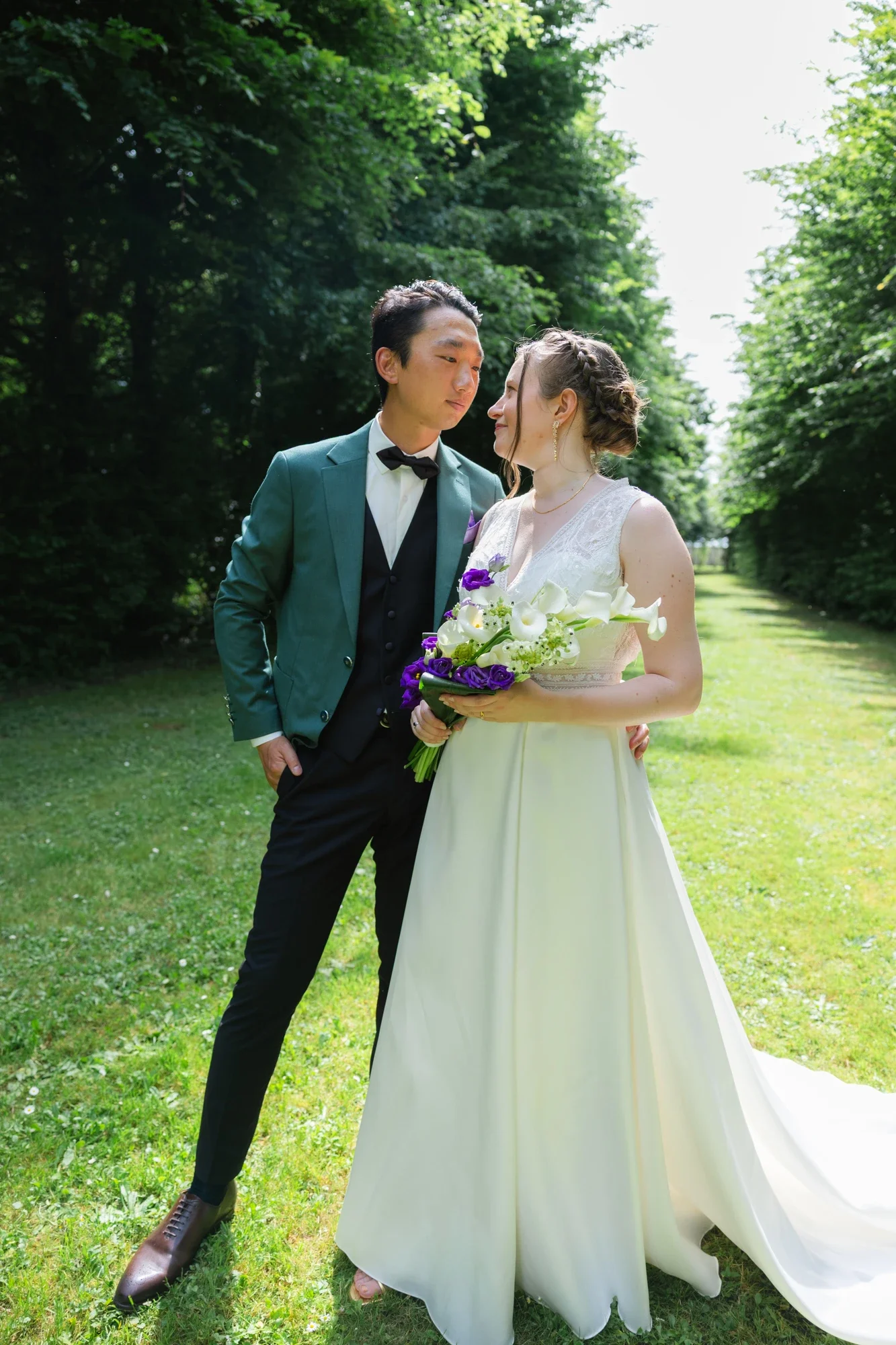 Un couple en mariage à l'extérieur dans un parc verdoyant, la femme tient un bouquet de fleurs blanches et violettes, ils se regardent tendrement.