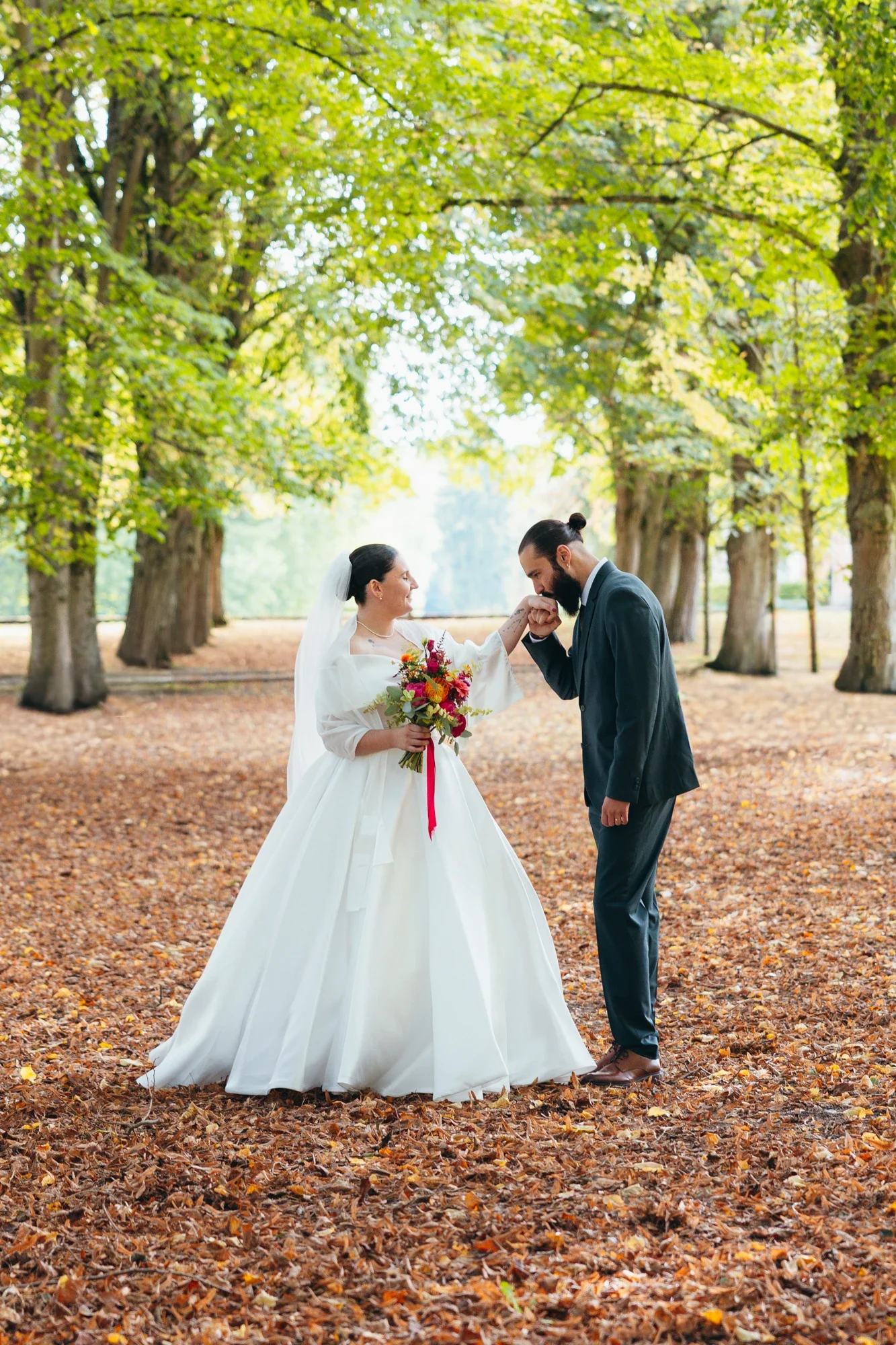 Un couple en mariage dans un parc entouré d'arbres verts, la femme en robe de mariée blanche et le homme en costume sombre, échangeant un bierge ou un geste affectueux.