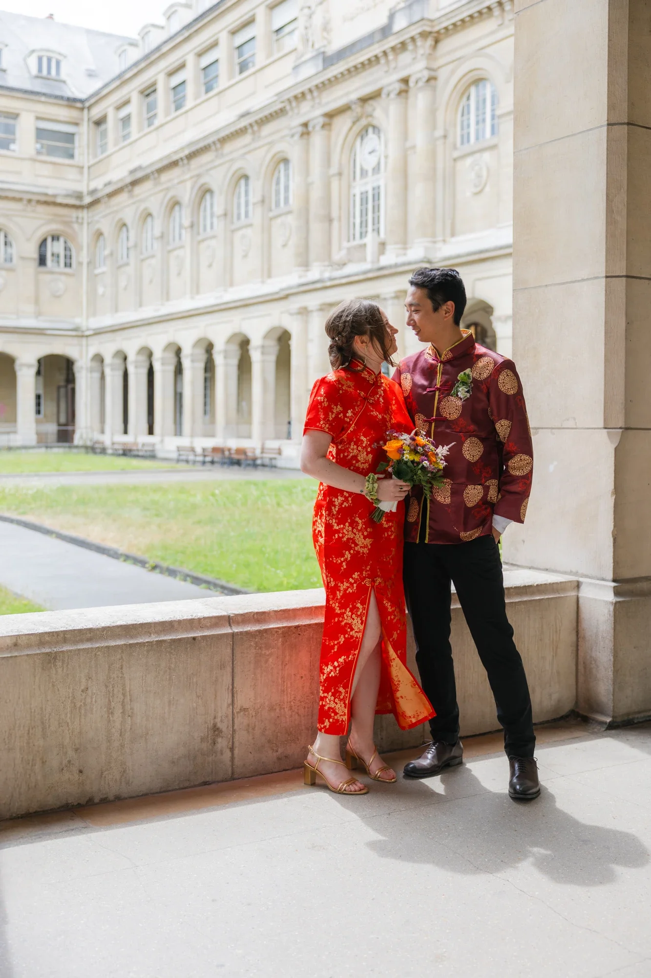 Un couple habillé en vêtements traditionnels chinois se regardant avec tendresse, debout dans un lieu architectural avec un bâtiment ancien en arrière-plan.