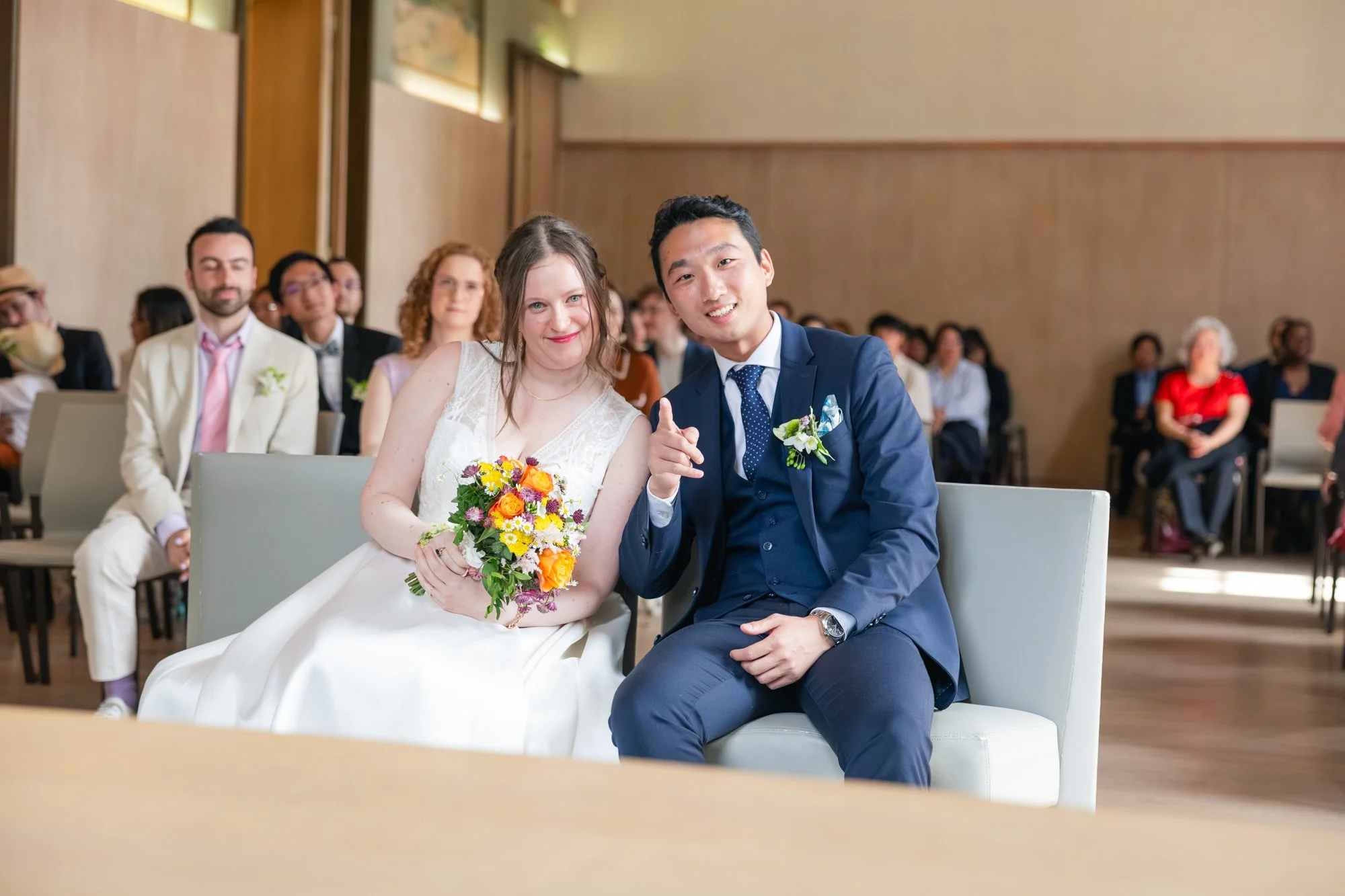 Un couple de mariés assis, entouré d'invités lors d'une cérémonie de mariage en intérieur.