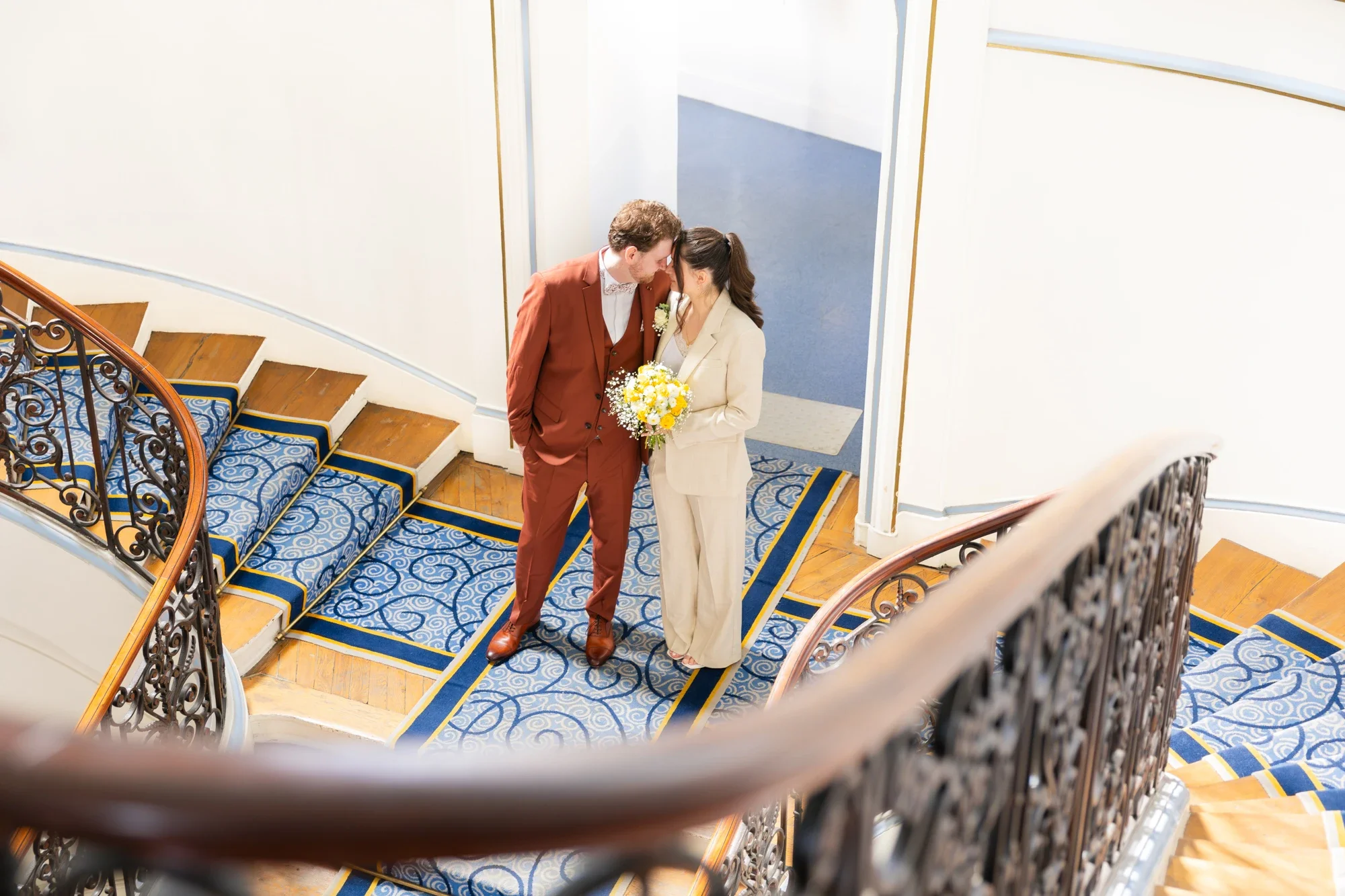Un couple en costume lors d'un mariage, se tenant dos à dos sur un escalier en bois recouvert d'un tapis bleu, dans une salle bien éclairée.